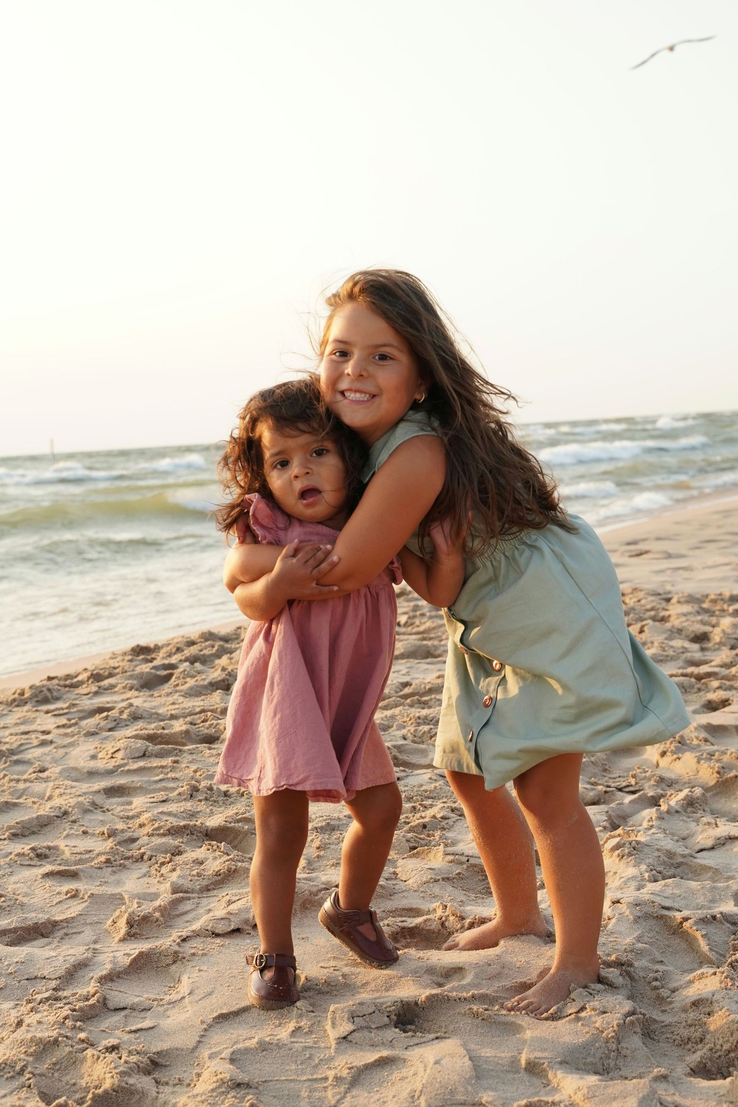 Two young sisters hugging and smiling on a beach at sunset, one in a pink dress and one in green.