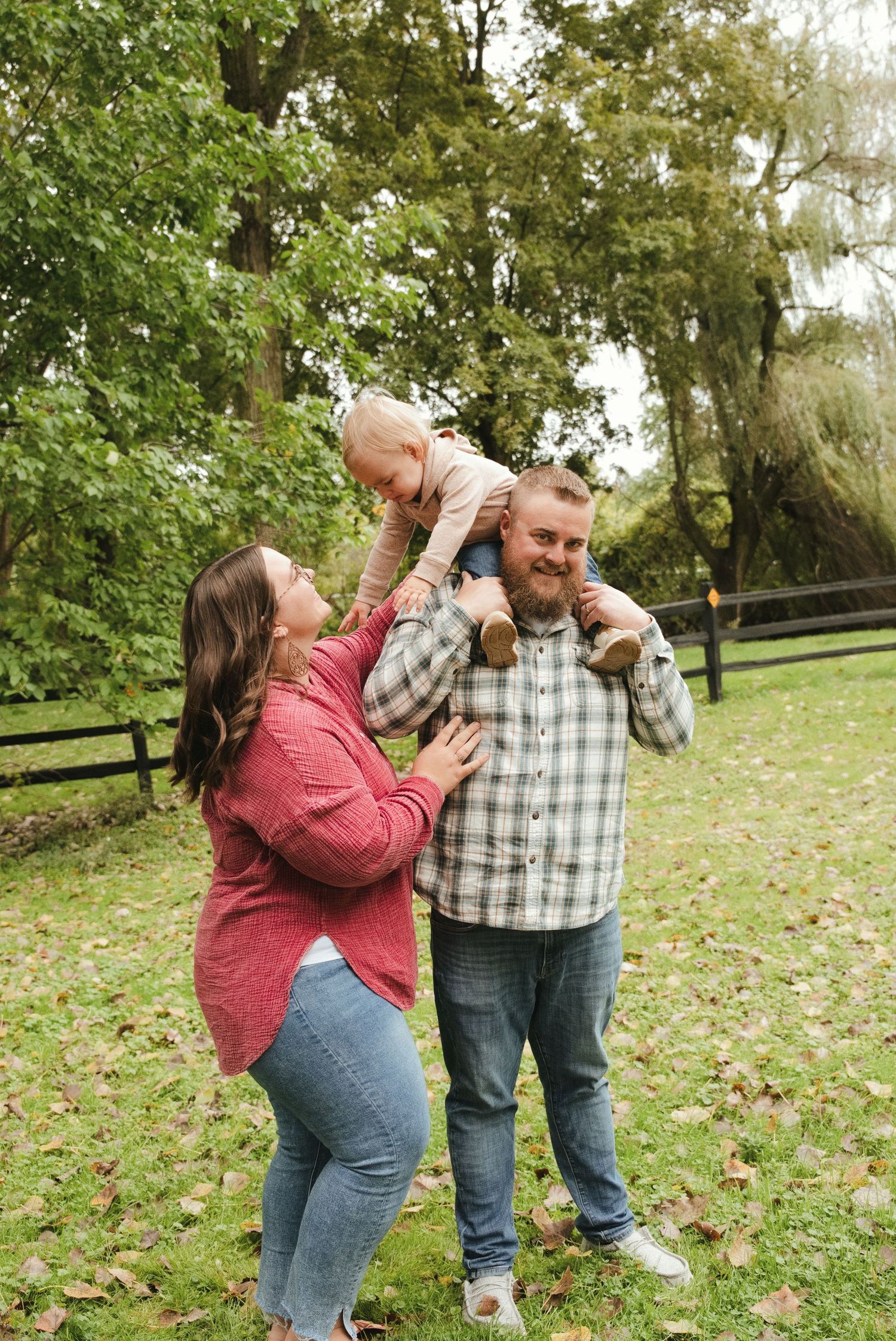 A smiling family of three poses outdoors on a grassy lawn as a toddler sits on dad's shoulders while mom looks up lovingly.