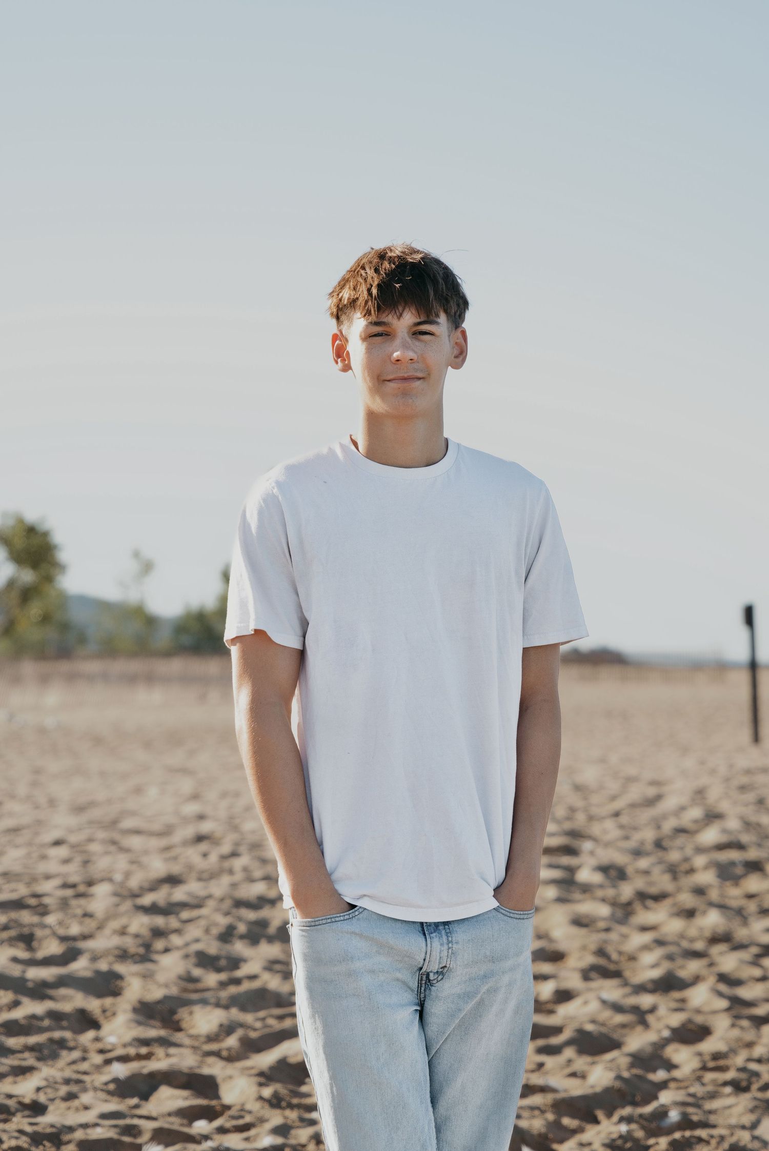 A teenage boy in a white t-shirt and jeans stands on a sandy beach, smiling at the camera on a sunny day.