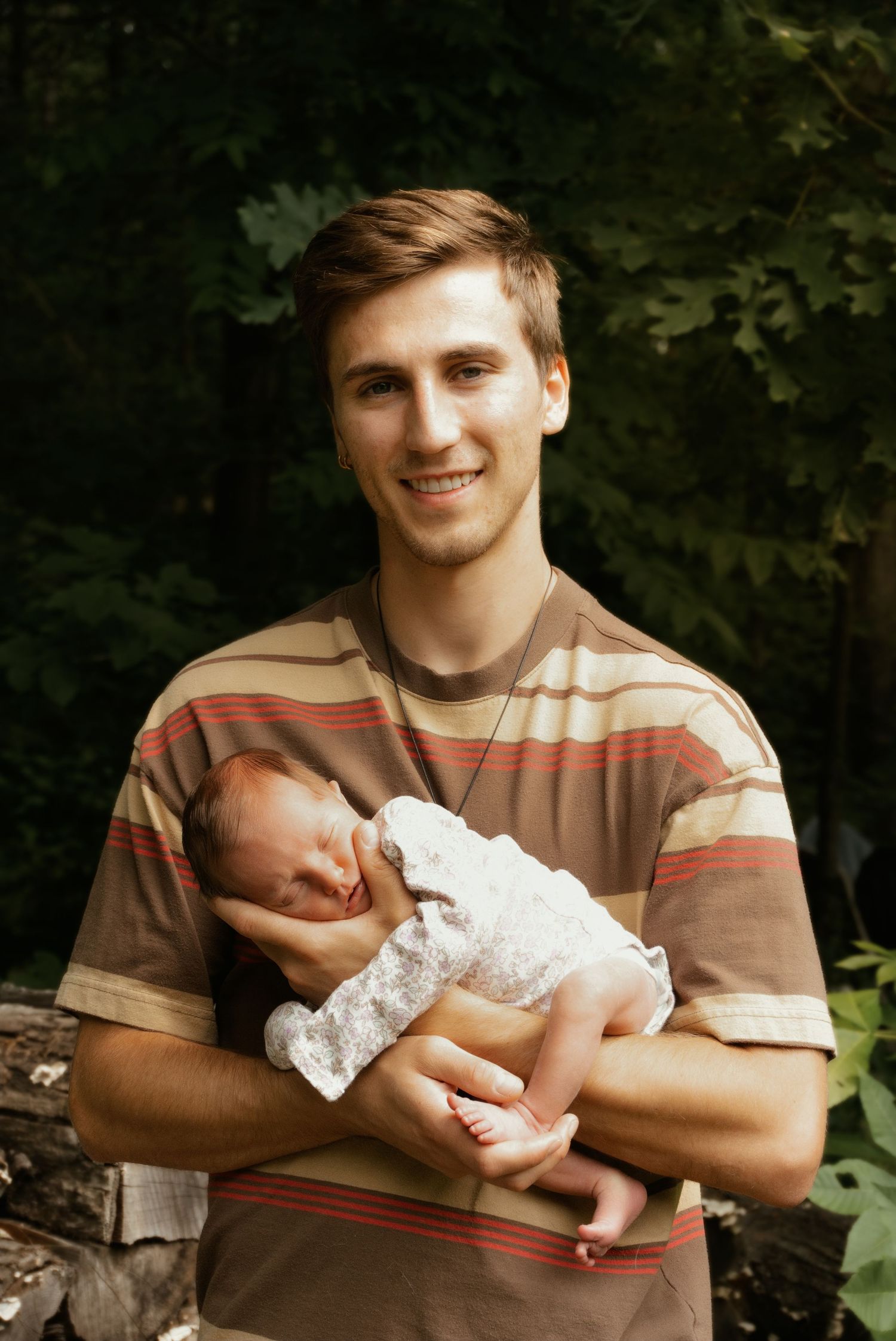 Smiling young father in a striped shirt proudly holds his sleeping newborn baby outdoors in nature.