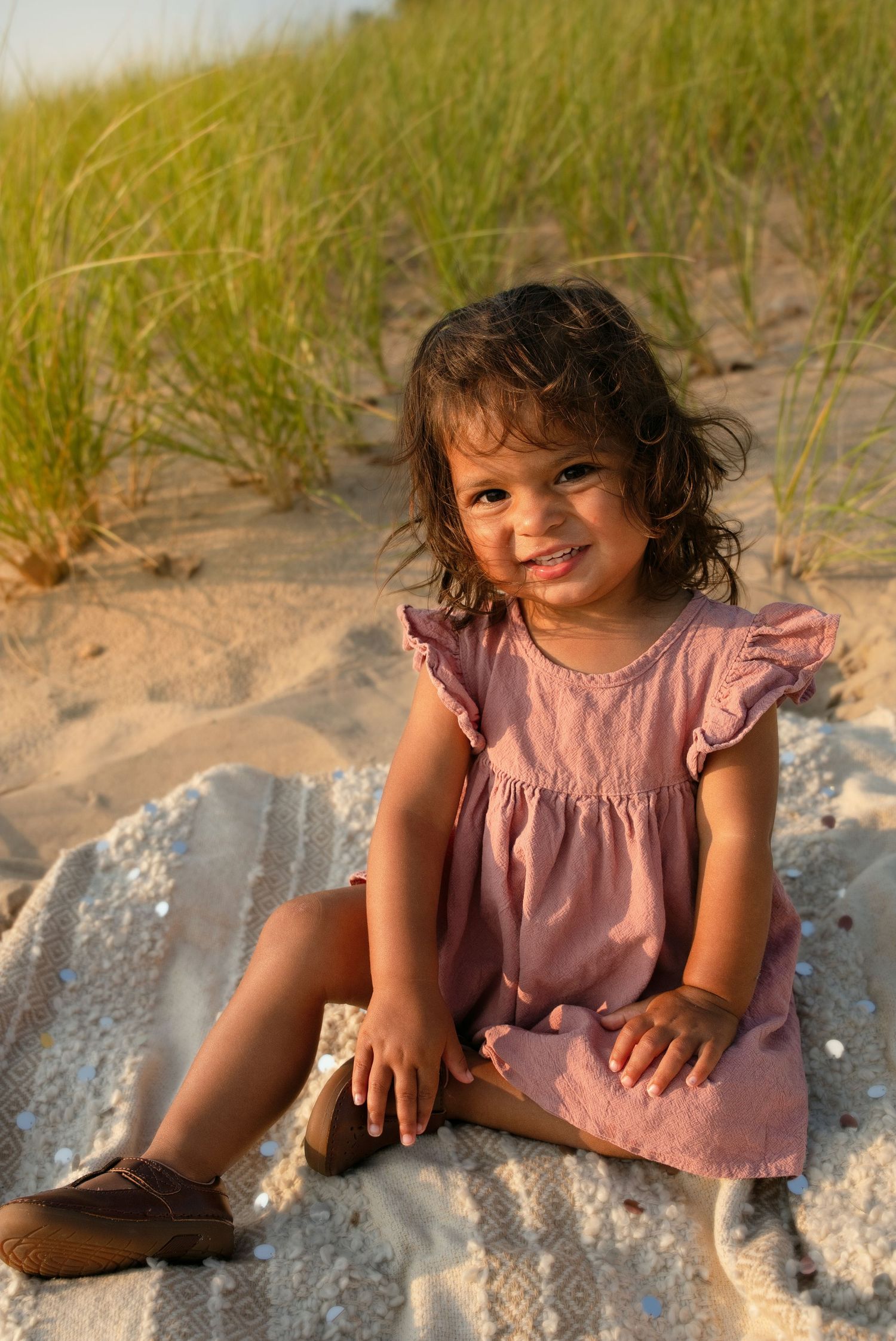 Toddler girl in pink dress sitting on blanket at sandy beach with grass dunes, smiling at camera.