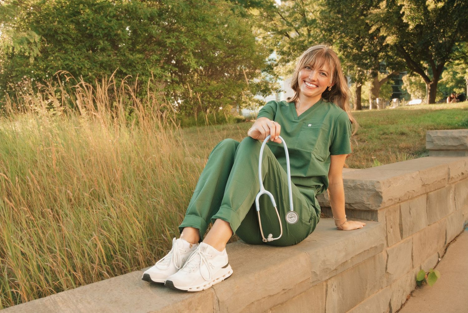 Female nurse in green scrubs sitting outdoors on stone steps, holding a stethoscope, smiling in a park setting.