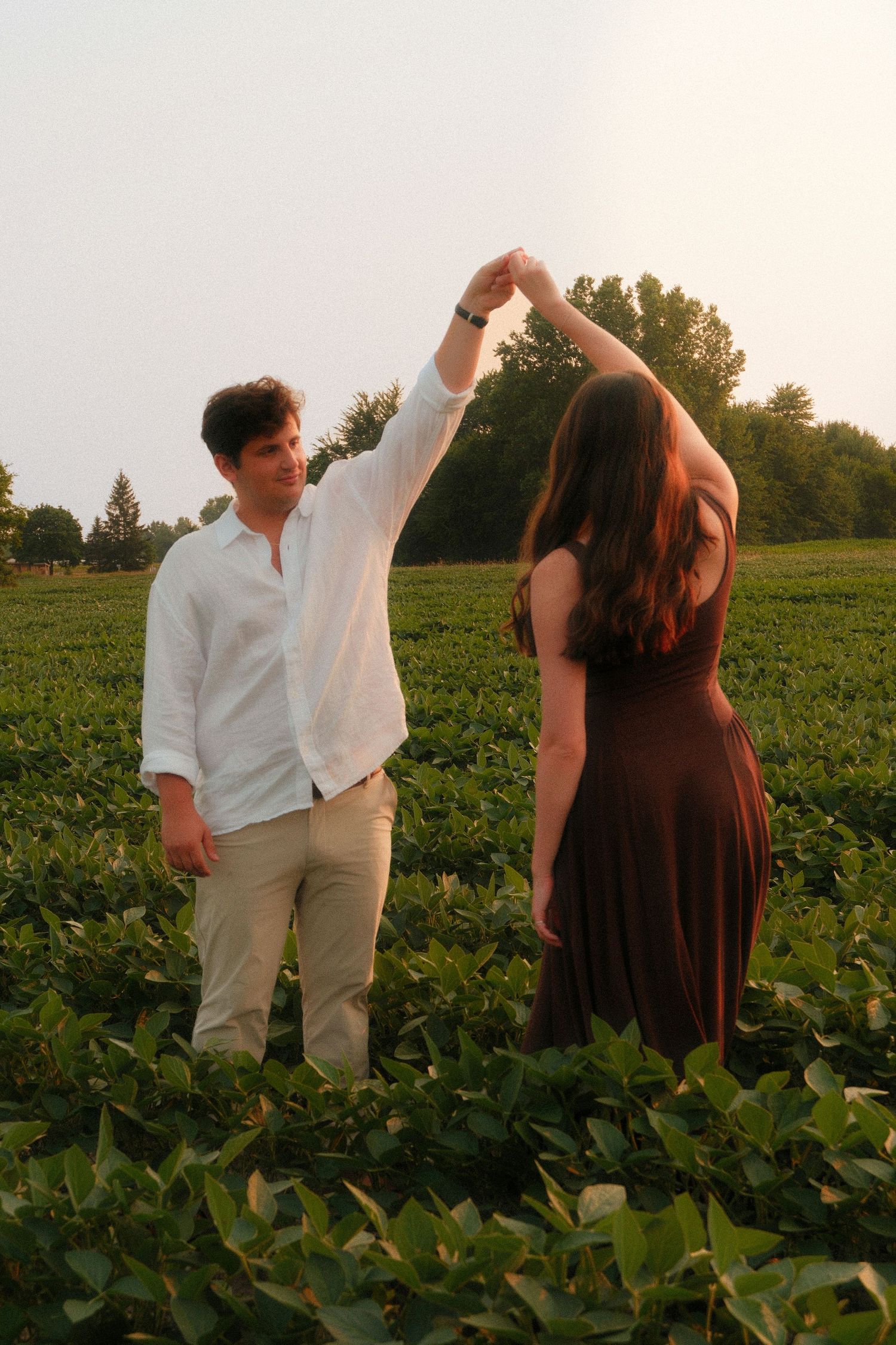 A couple dances together in a lush green field at golden hour, man in white shirt twirling woman in brown dress.