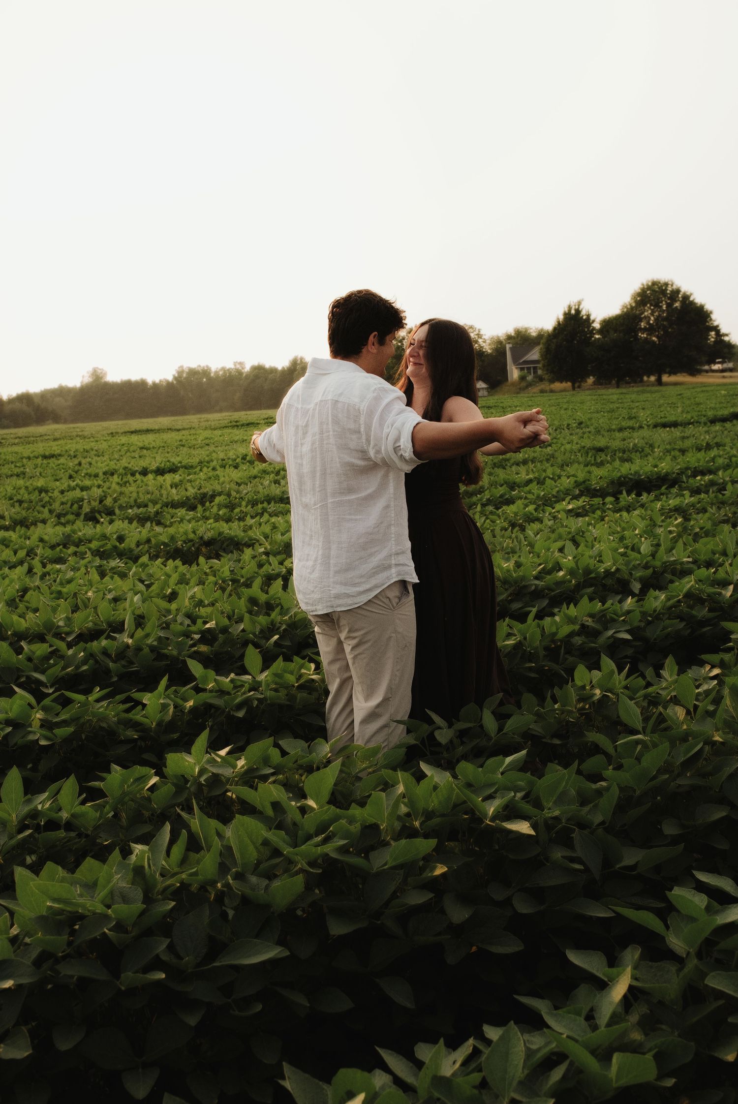 Couple embracing in a lush green farm field at sunset, woman in black dress and man in white shirt.