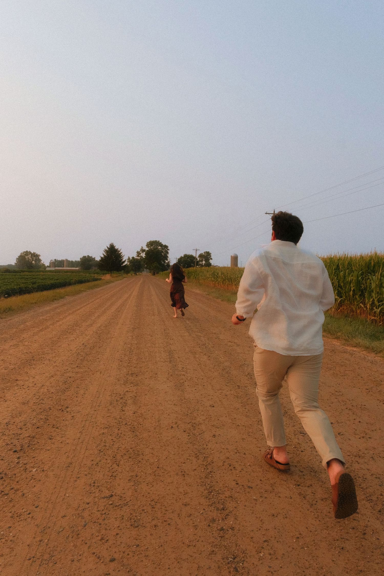 Couple running down a rural dirt road at sunset with cornfields and trees on either side.