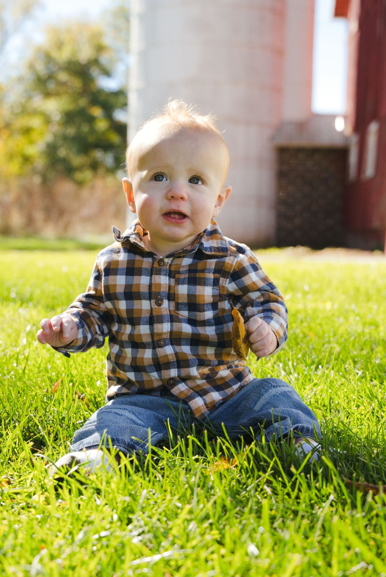 Smiling baby in a plaid flannel shirt and jeans sits on bright green grass in front of a red barn and silo on a sunny day.