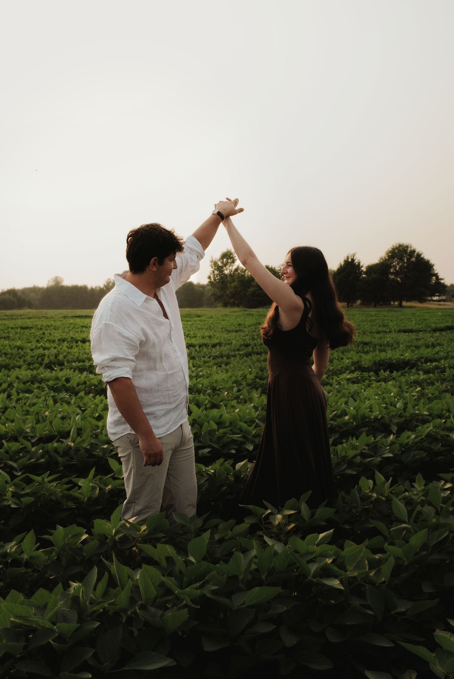 Romantic couple dancing and spinning hands in a lush green farm field at sunset.