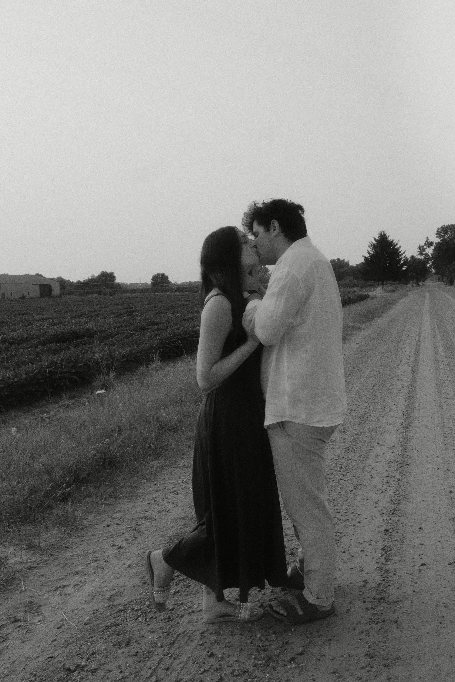 Black and white photo of romantic couple embracing on a rural dirt road surrounded by open fields.