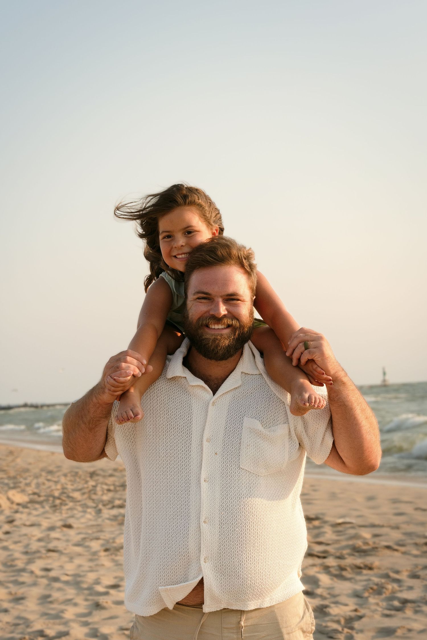 Smiling bearded man carrying young girl on shoulders at beach during golden hour.