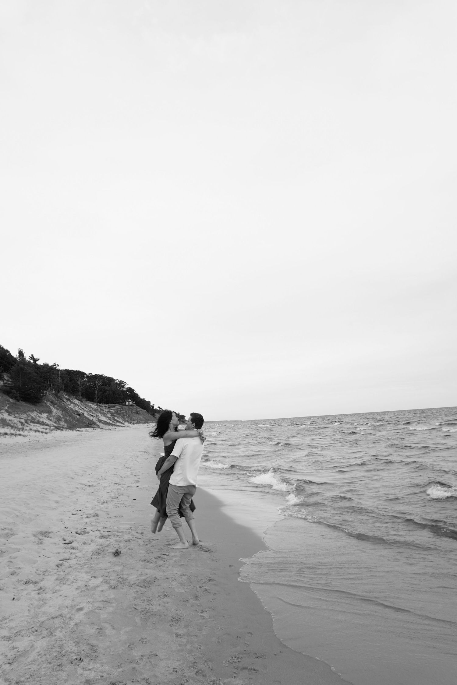 Black and white photo of a couple embracing on a sandy beach with ocean waves and tree-lined dunes in the background.