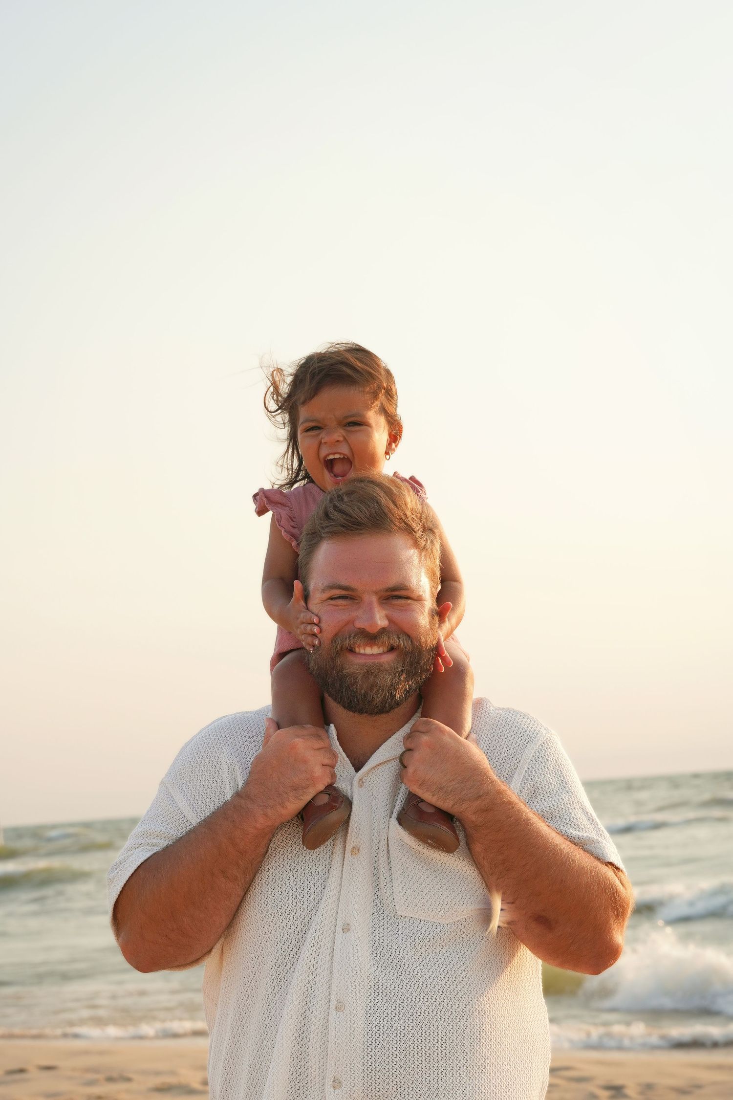 Happy bearded father with laughing toddler girl on shoulders at sunny beach.