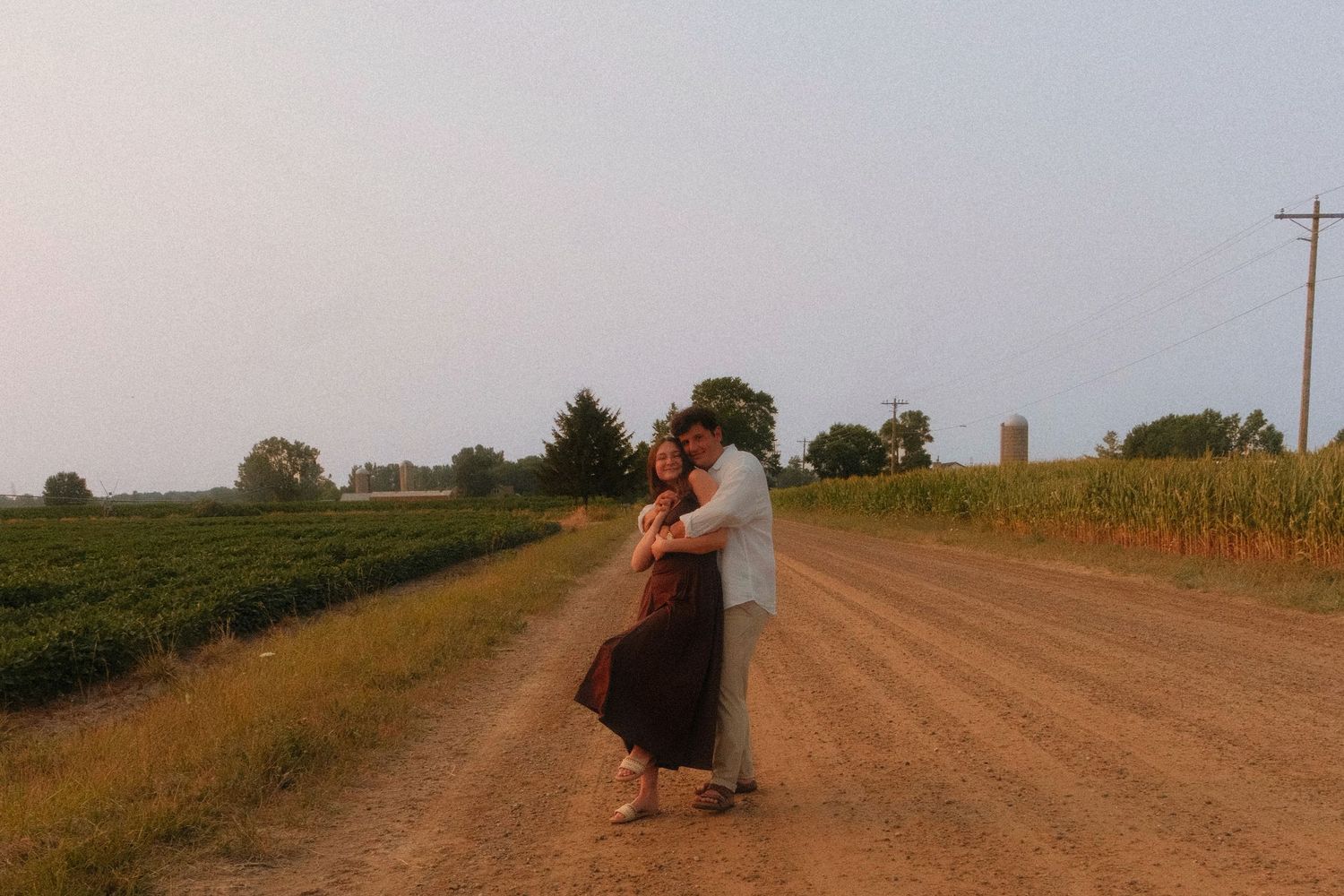 Couple embracing on a rural dirt road surrounded by green fields and corn crops during golden hour.