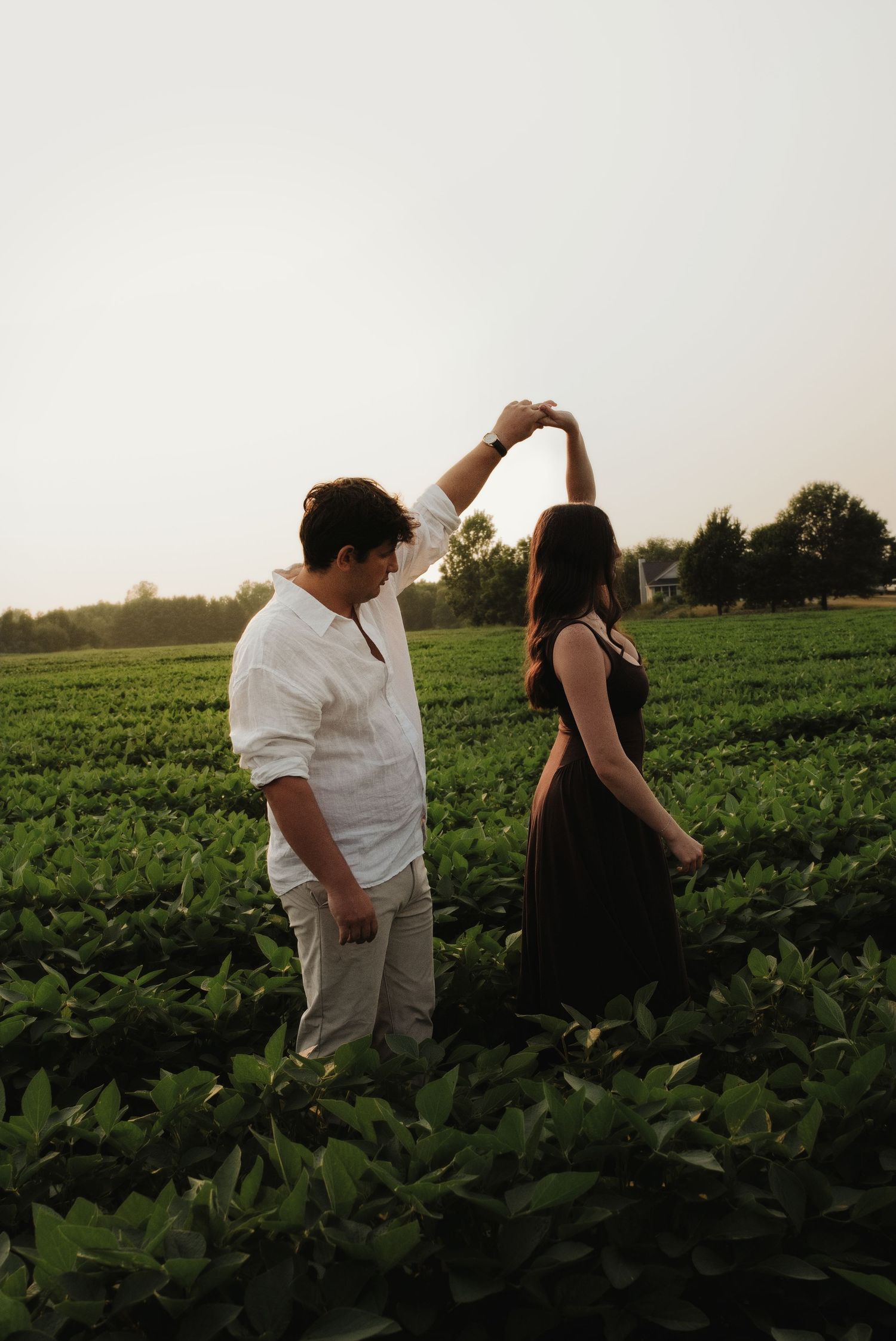 Couple dancing playfully in a lush green crop field during a warm summer sunset.