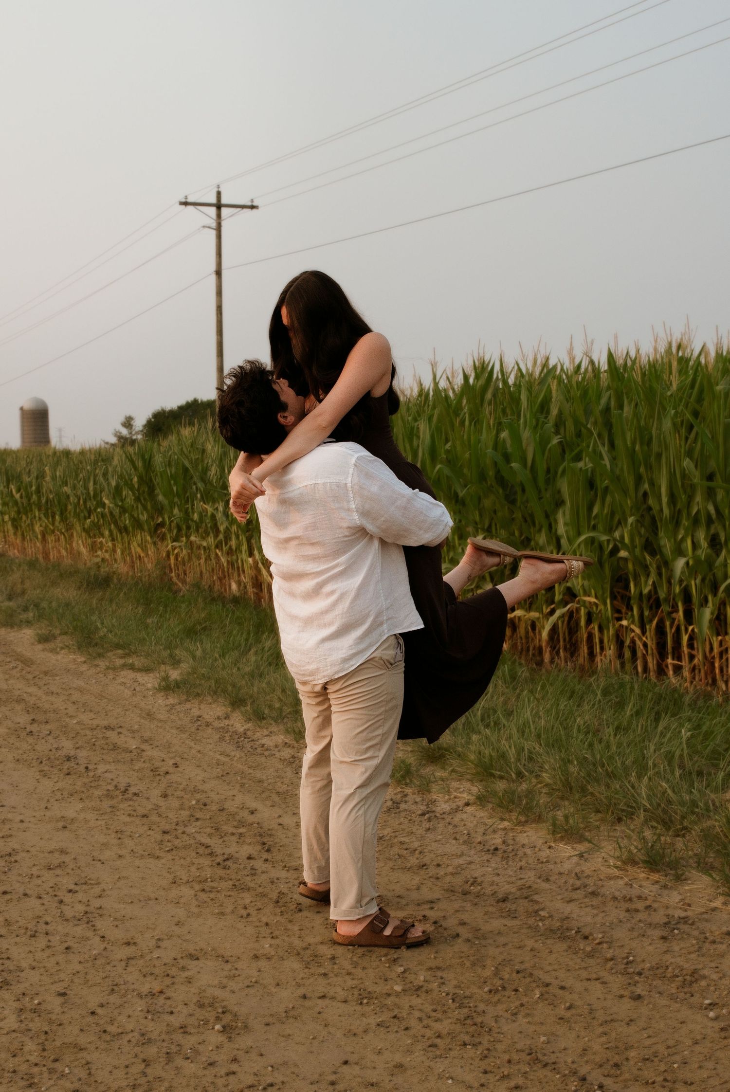 Man lifting woman in his arms on a dirt path beside a tall cornfield at dusk.