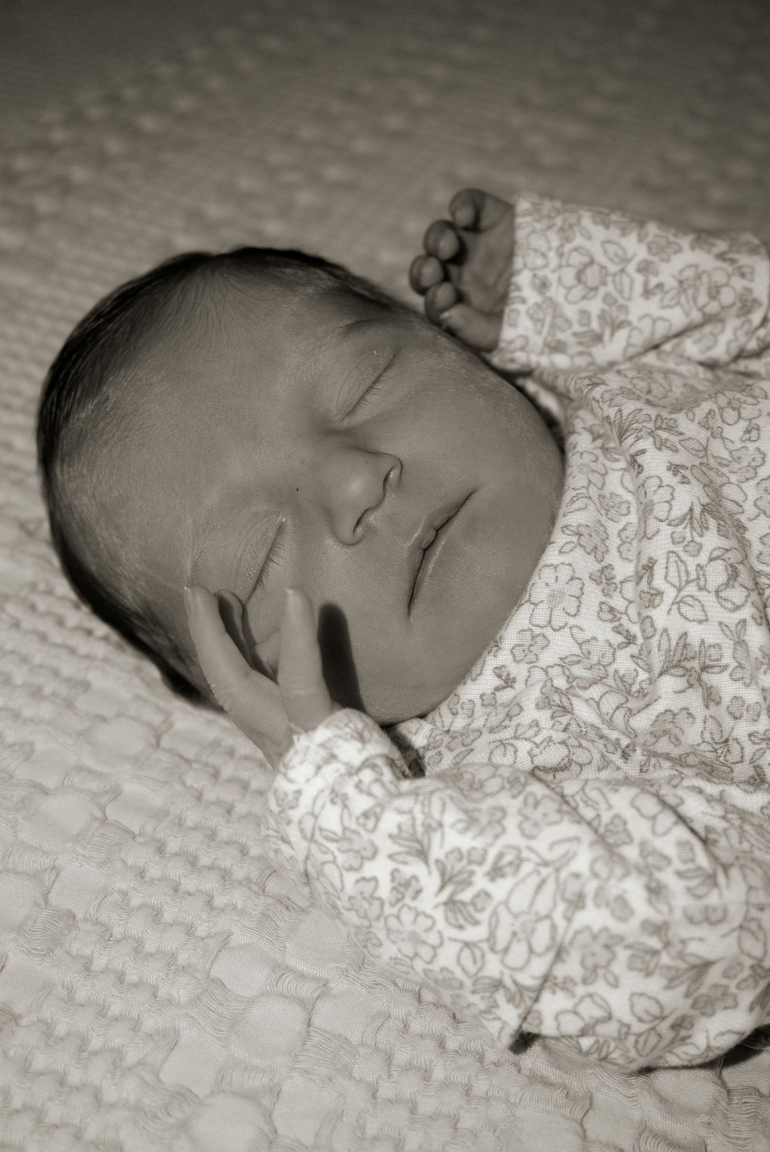 Black and white photo of a peaceful sleeping newborn baby in a floral onesie with tiny hand near face.