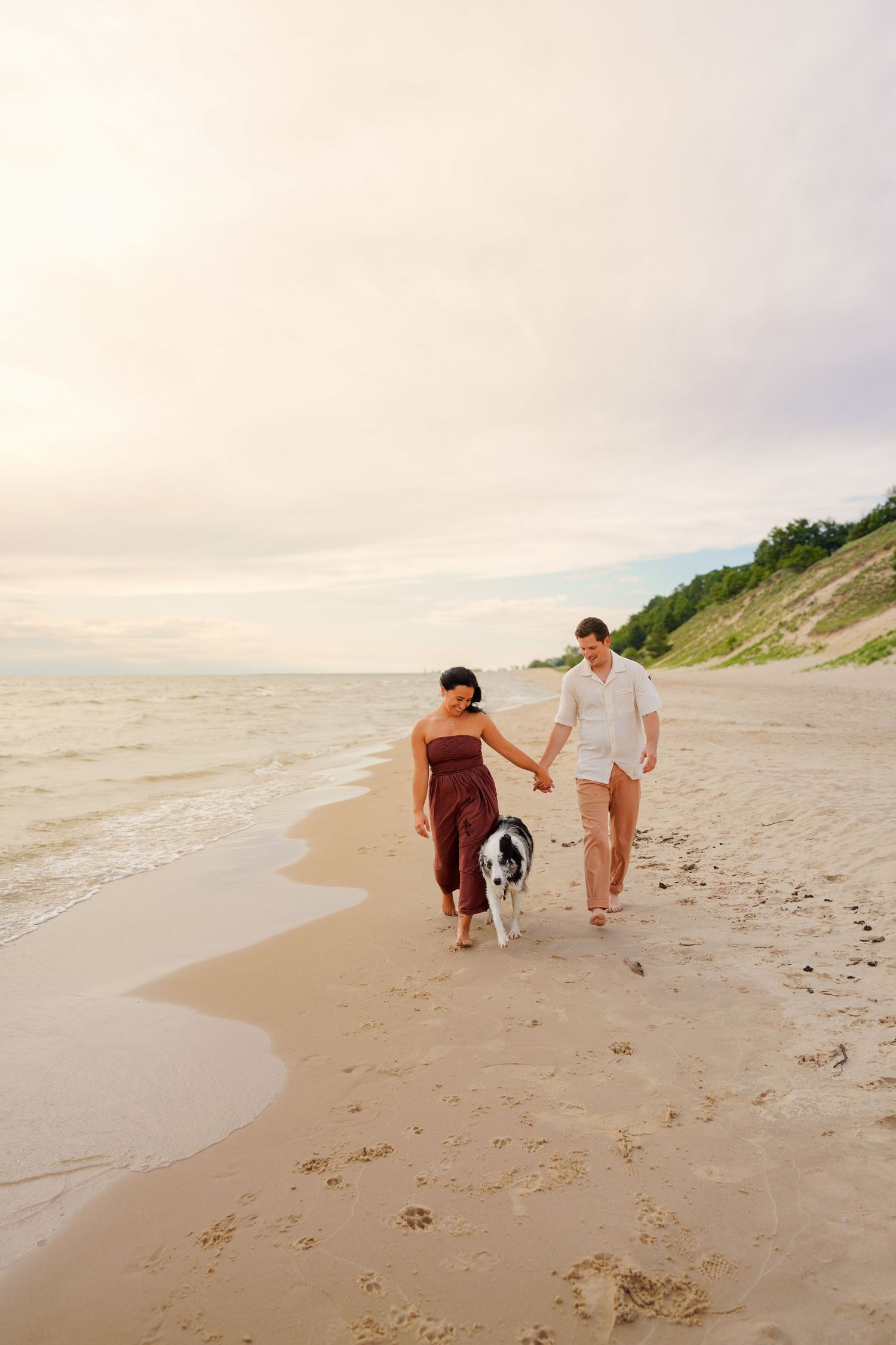 Couple holding hands walking their black and white dog along a sandy beach at sunset.