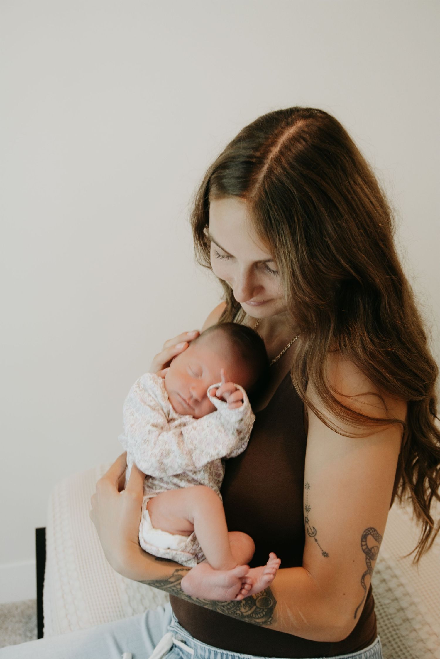 Tattooed mother tenderly holding sleeping newborn baby against her chest in a soft, natural light setting.