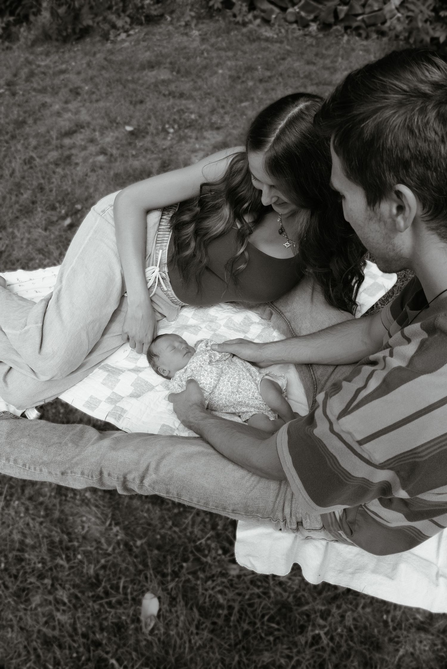 Black and white aerial view of new parents on a quilt in grass, tenderly looking at their newborn baby.
