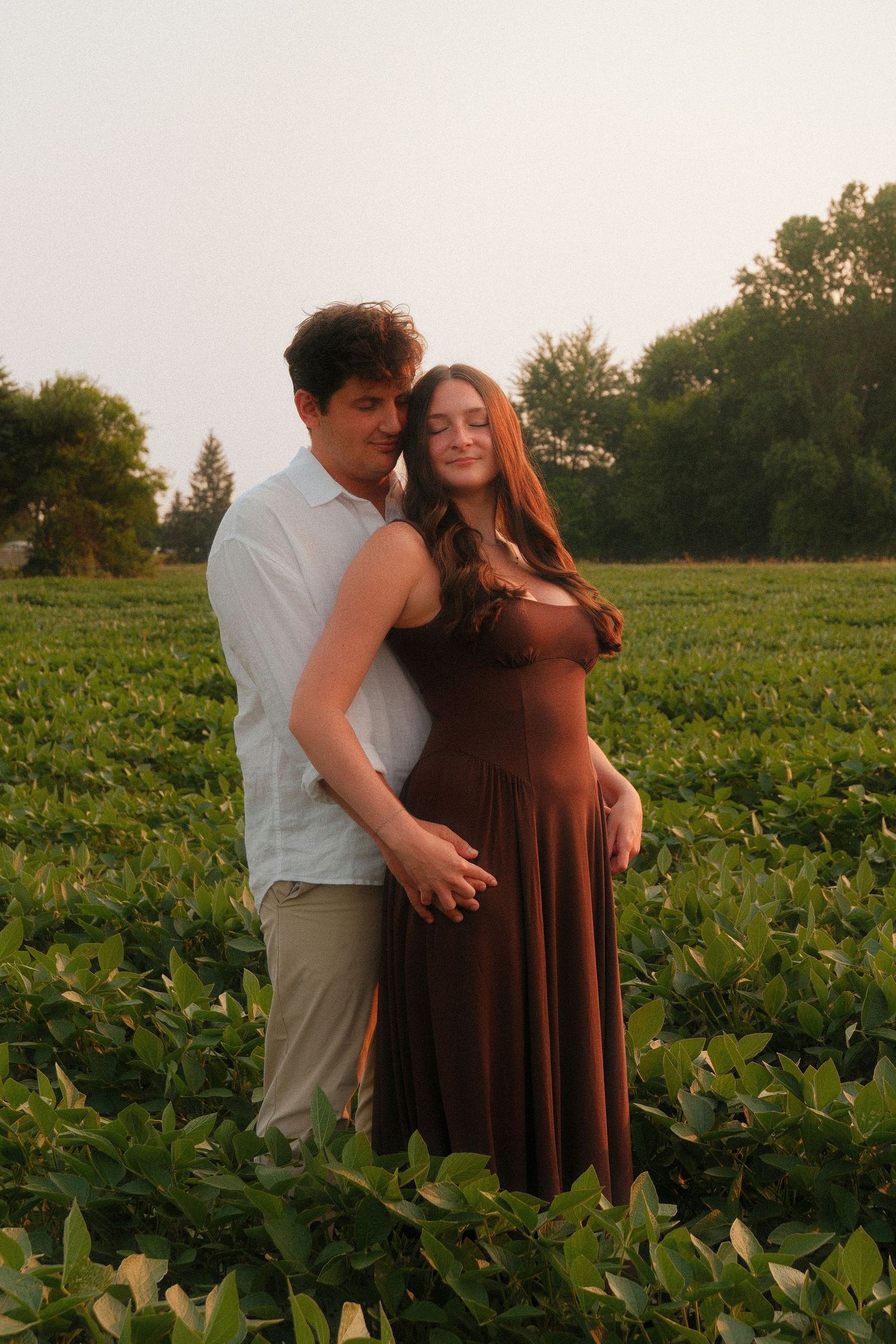 Couple embracing in a lush green field at golden hour, woman in brown dress holding her baby bump.