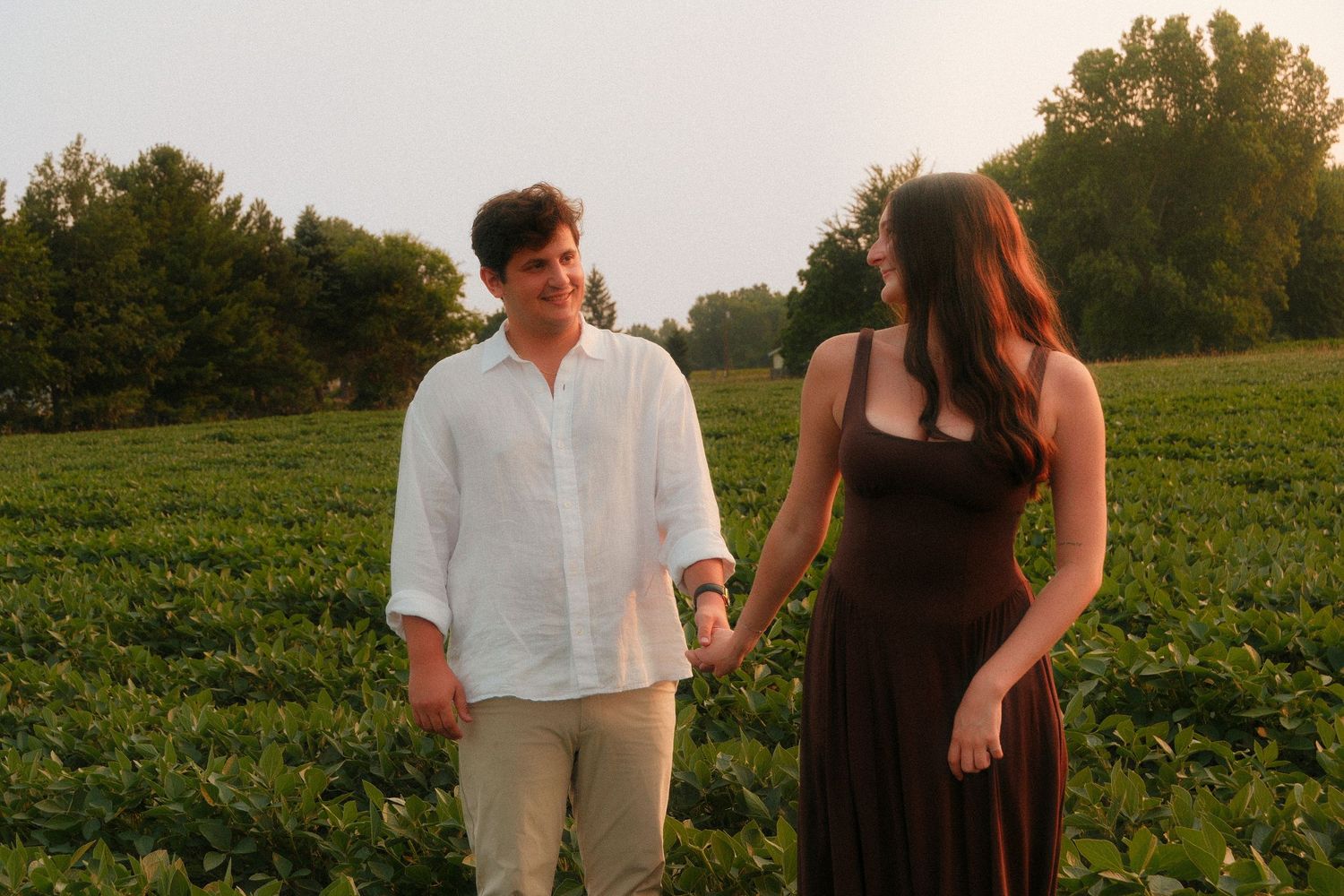 Couple holding hands in a lush green field at sunset, man in white linen shirt, woman in black dress.