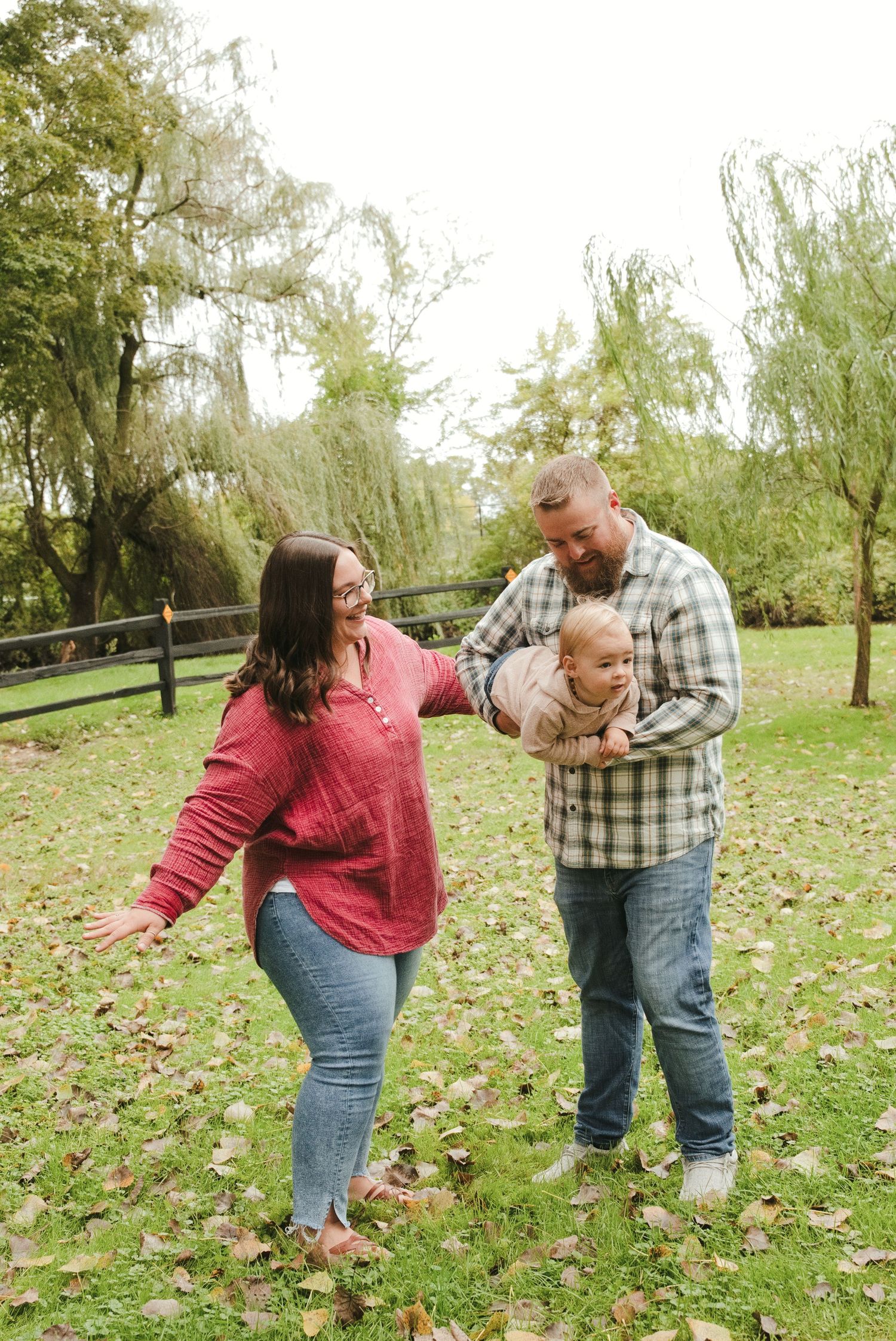 Parents play with their baby on a leaf-covered lawn near a wooden fence during an autumn family photo session.