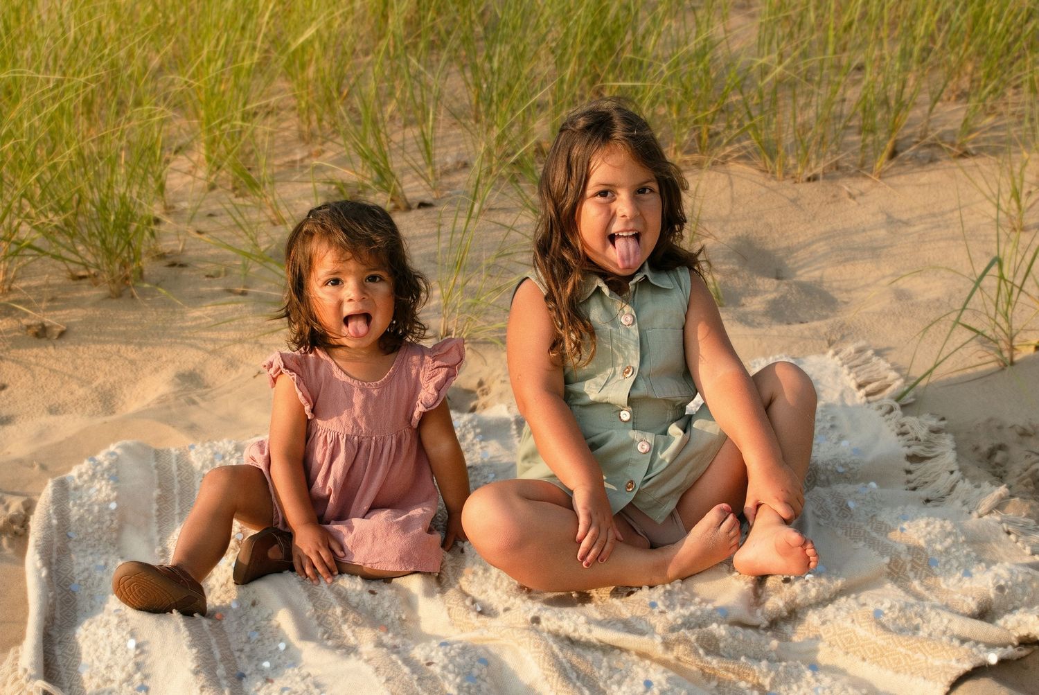 Two young girls sitting on a blanket at the beach, smiling and sticking out their tongues near sand dunes.
