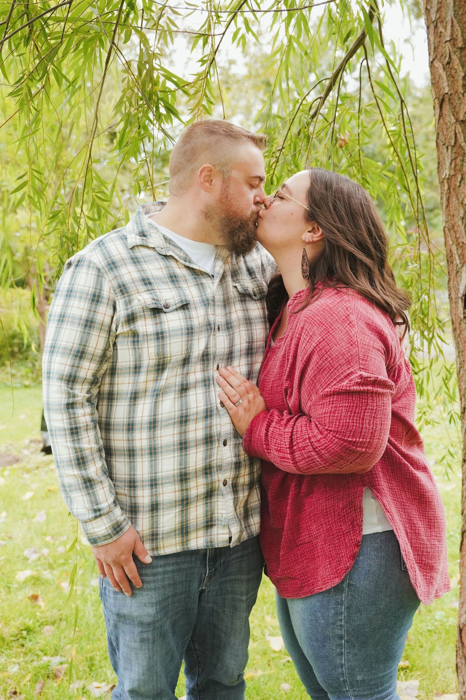 Couple kissing under a willow tree in a park, he wears a plaid flannel shirt and she a red cardigan, during an engagement photo session.