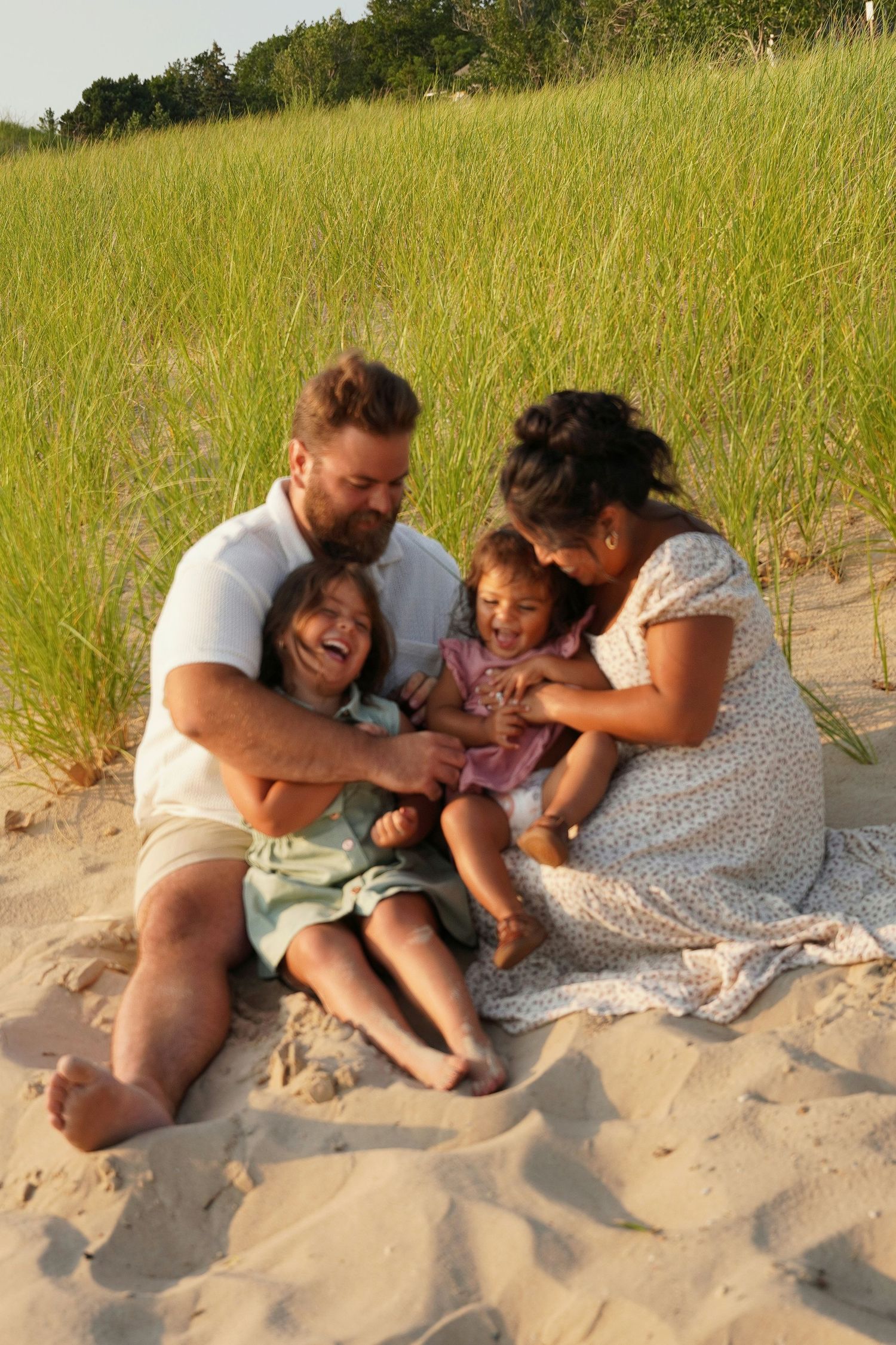 Happy family of four laughing together on sandy beach dunes with green sea grass in warm golden sunlight.
