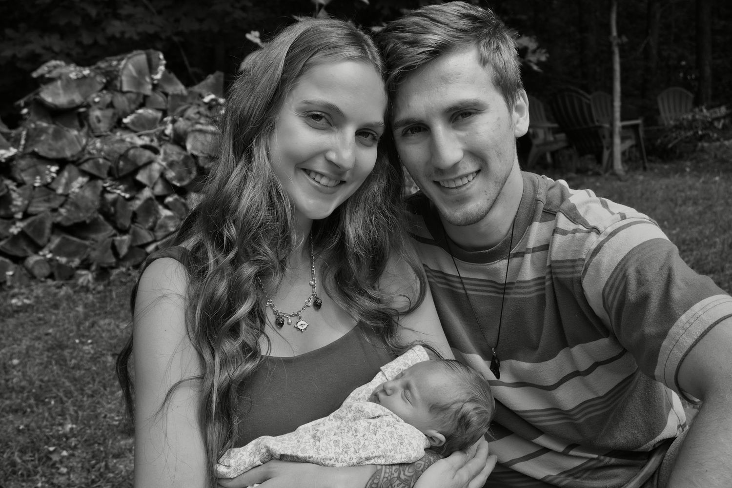 Black and white photo of smiling couple holding newborn baby outdoors near stacked firewood.