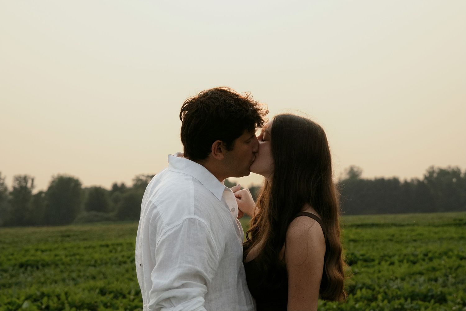 A romantic couple shares a tender kiss outdoors with lush green fields and a soft hazy sky in the background.