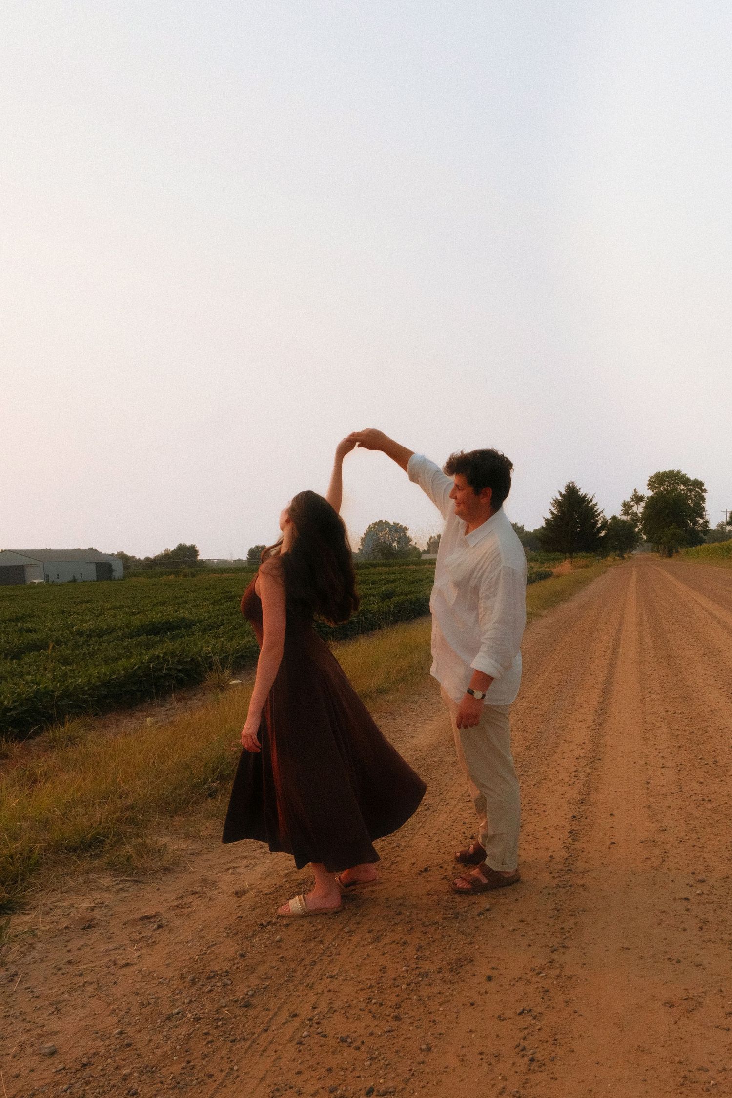 Couple dancing on a dirt country road at sunset, man spinning woman in a flowing brown dress.