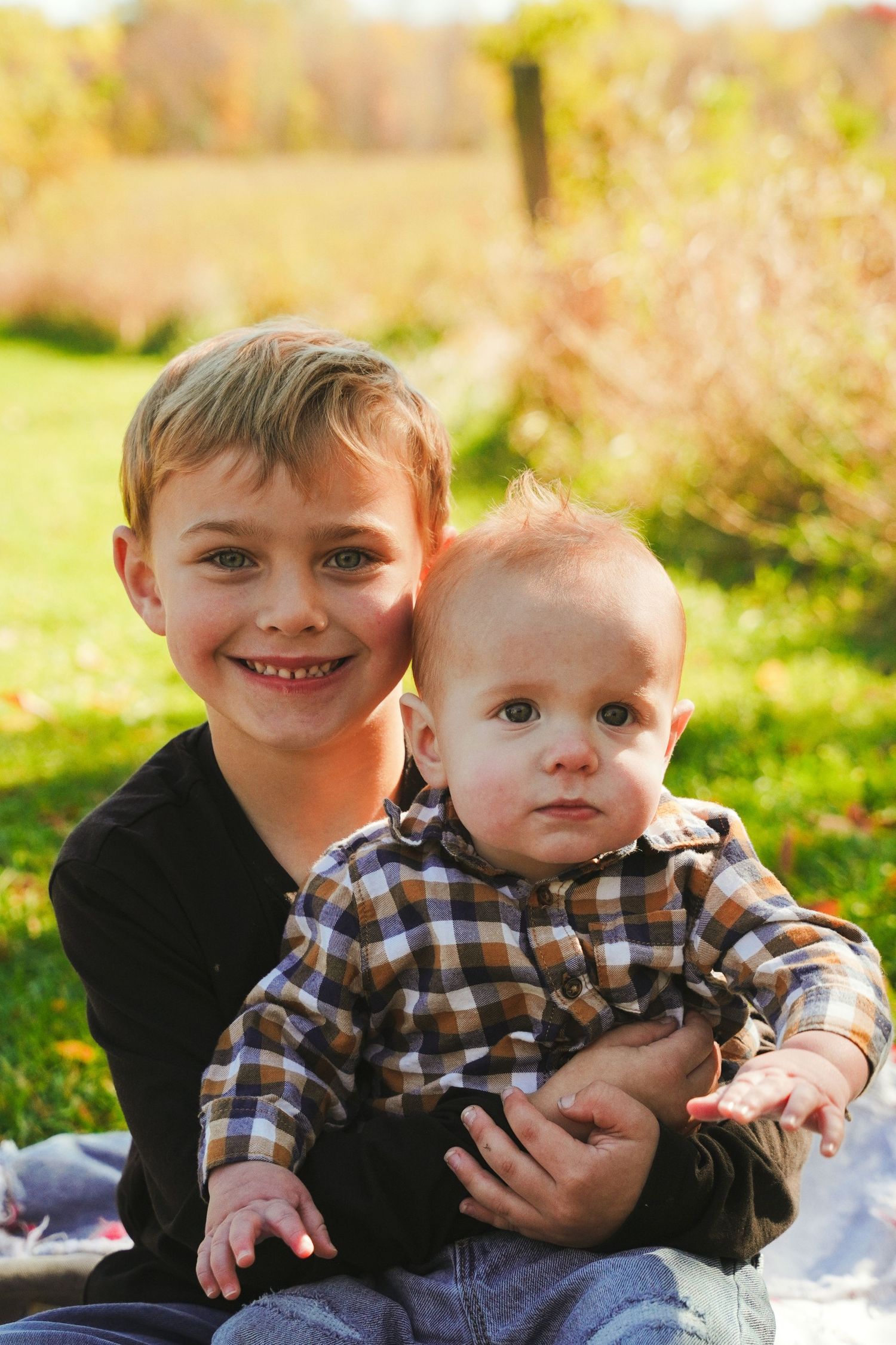 Smiling older boy in a black shirt holds a baby in a plaid flannel shirt while sitting on grass in a sunny autumn field.