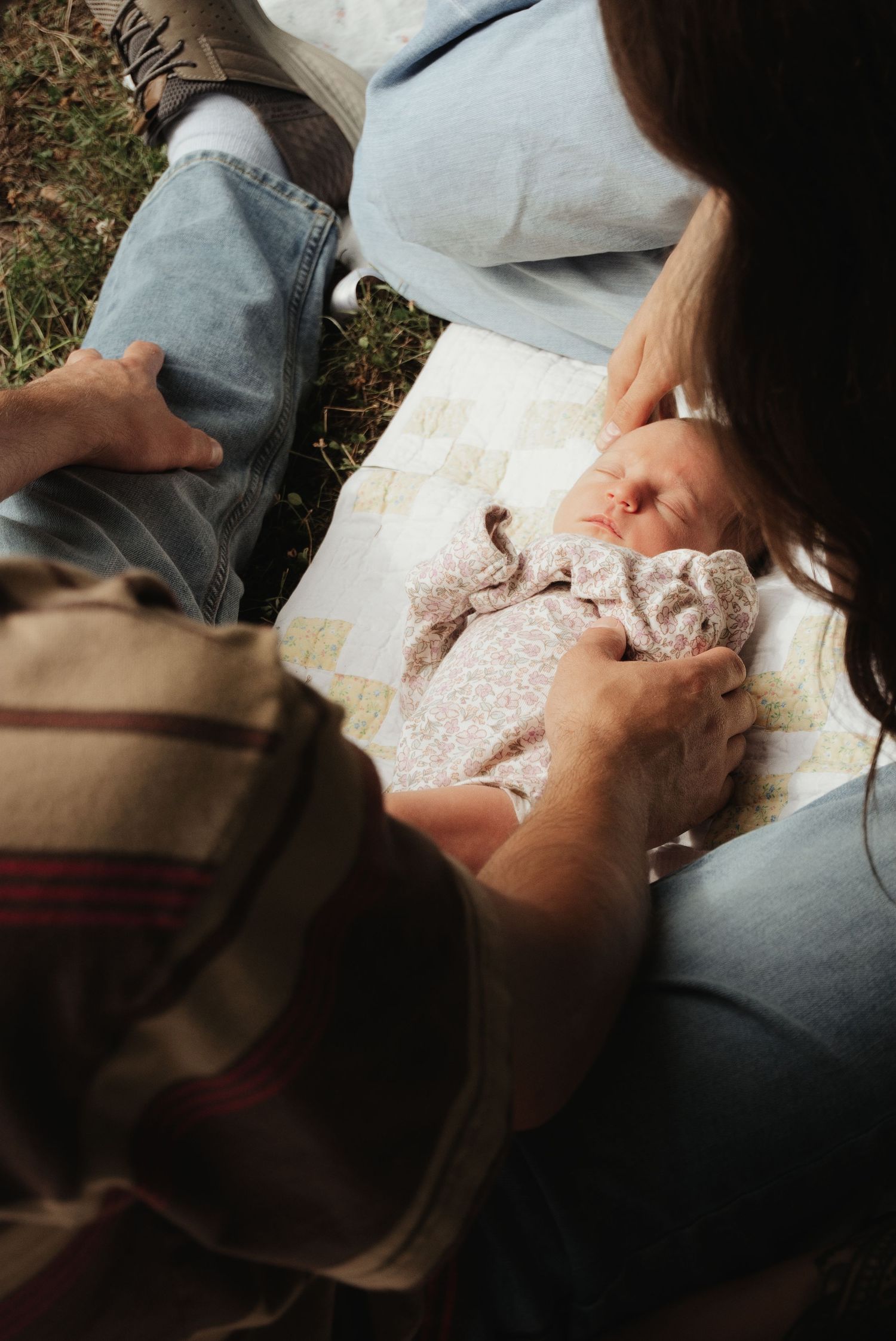 Two parents sitting outdoors on a quilt, lovingly gazing down at their peaceful sleeping newborn baby.
