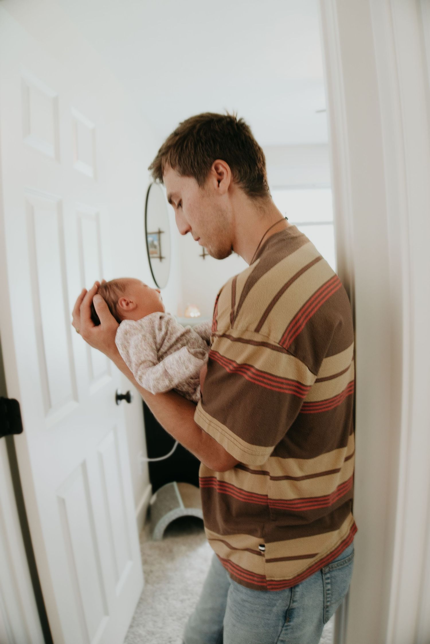 Young father in striped shirt tenderly holds newborn baby while standing in a bright, modern bathroom doorway.