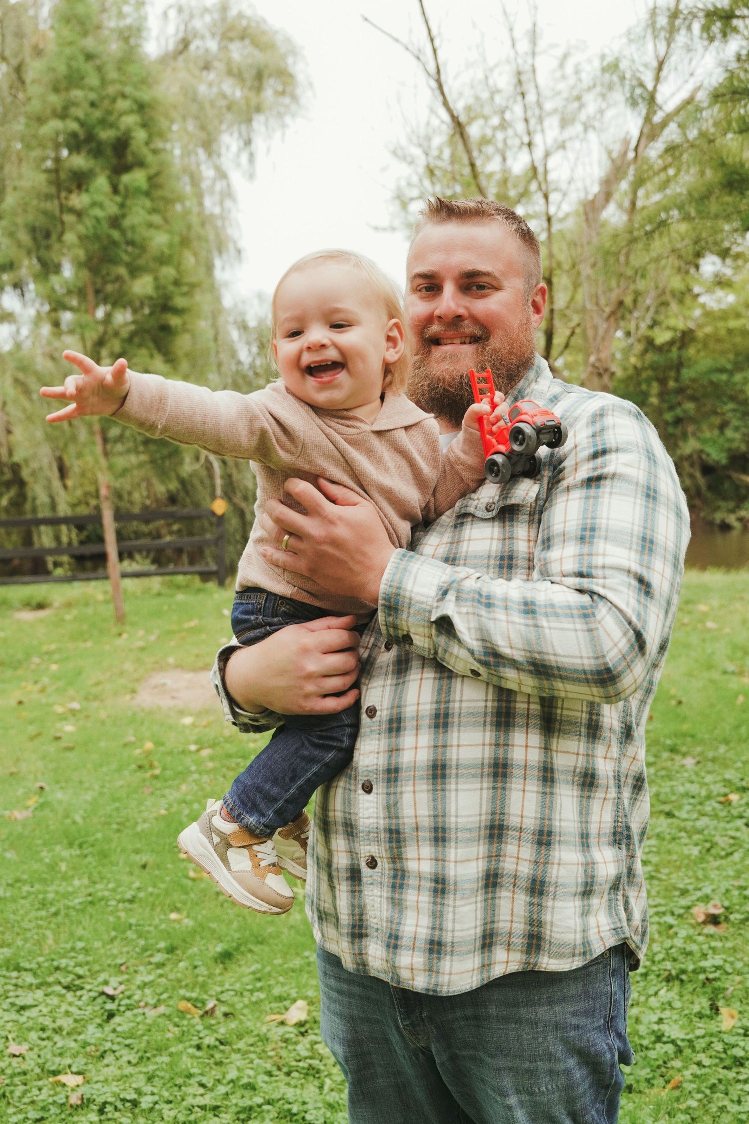 Bearded dad in a plaid shirt holds his giggling toddler, who reaches an arm out, in a grassy park during autumn.