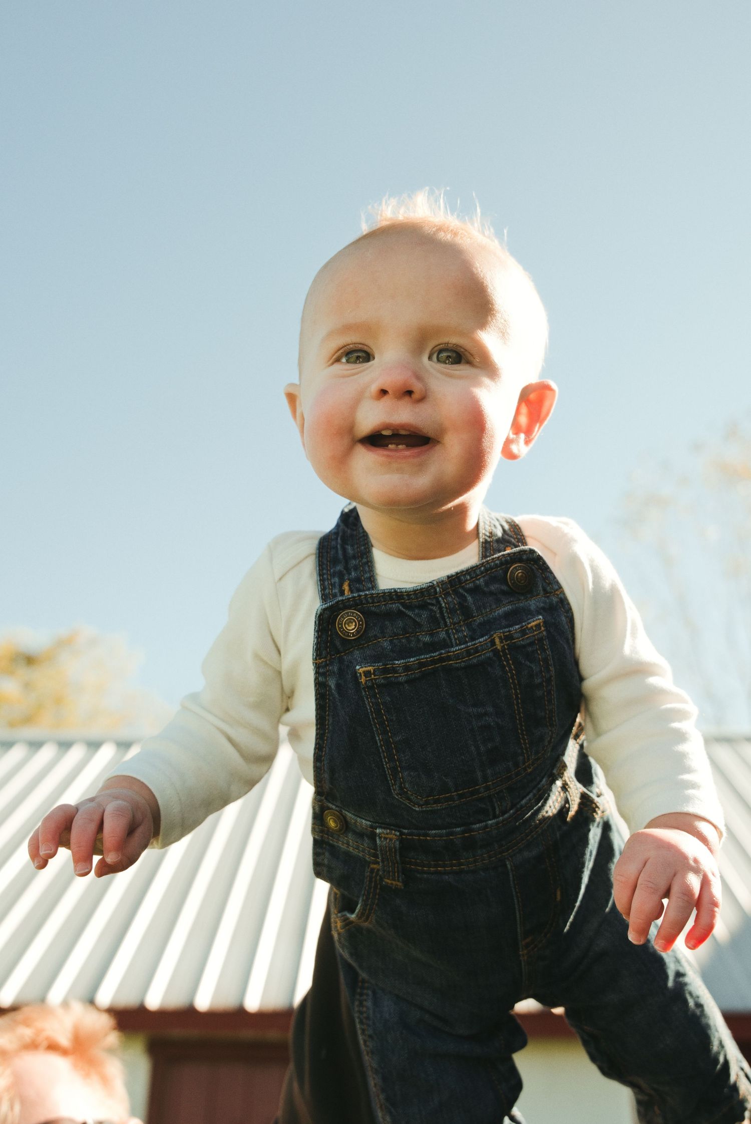 Smiling baby in denim overalls and a white long-sleeve shirt is held up against a clear blue sky near a metal-roofed building.