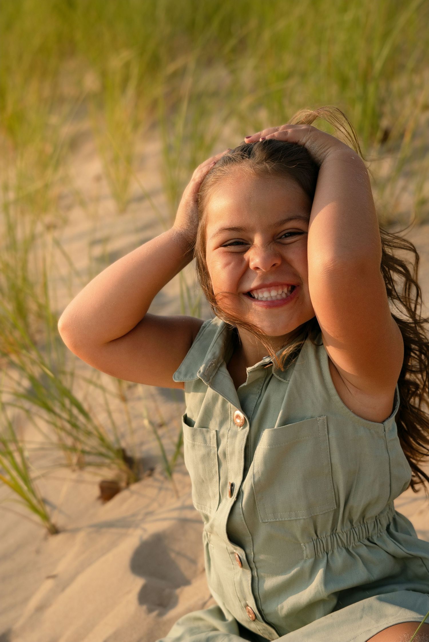 A young girl smiles joyfully on sandy beach dunes, hands in hair, wearing a sage green dress in golden sunlight.