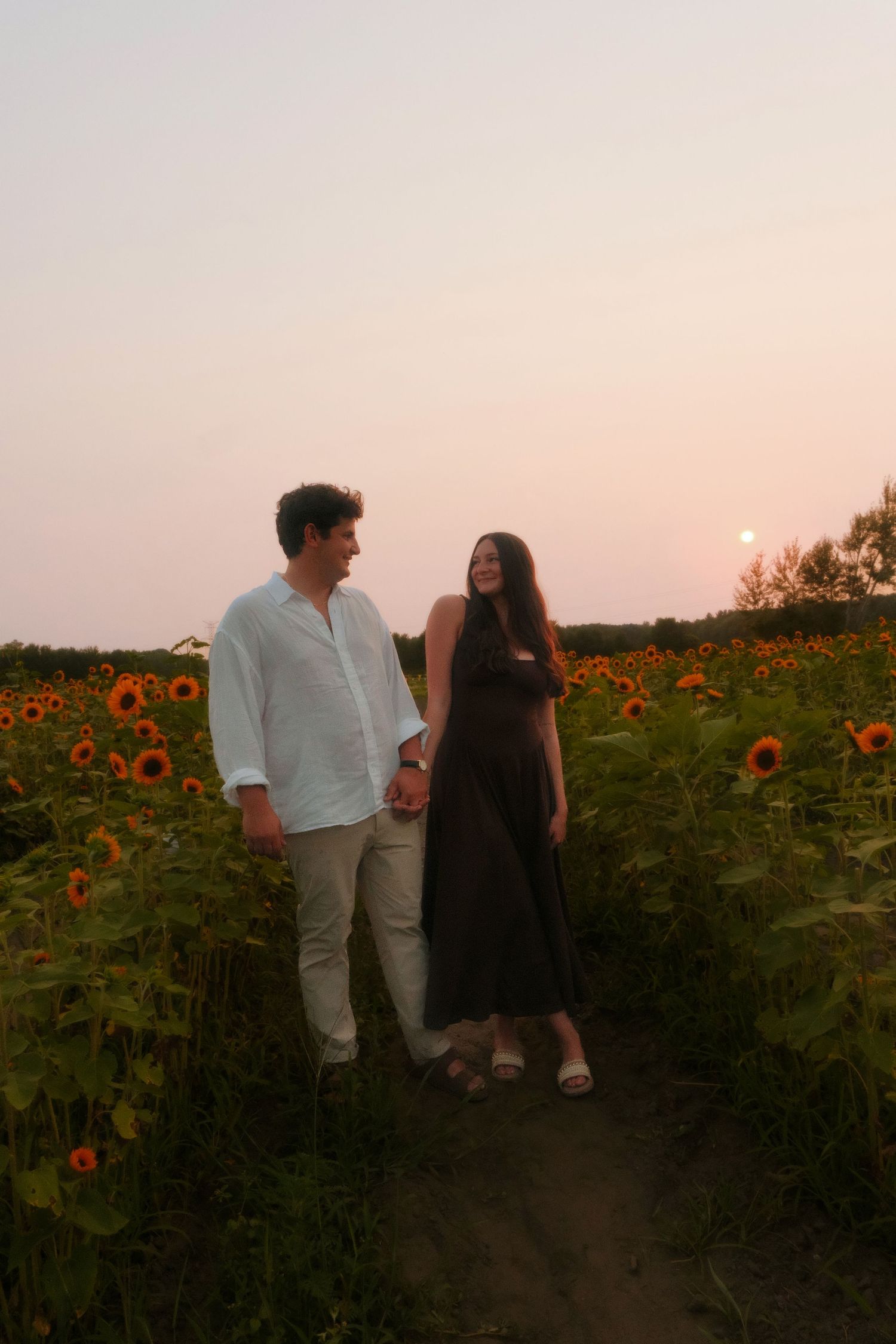 Couple holding hands on a dirt path through a sunflower field at sunset.