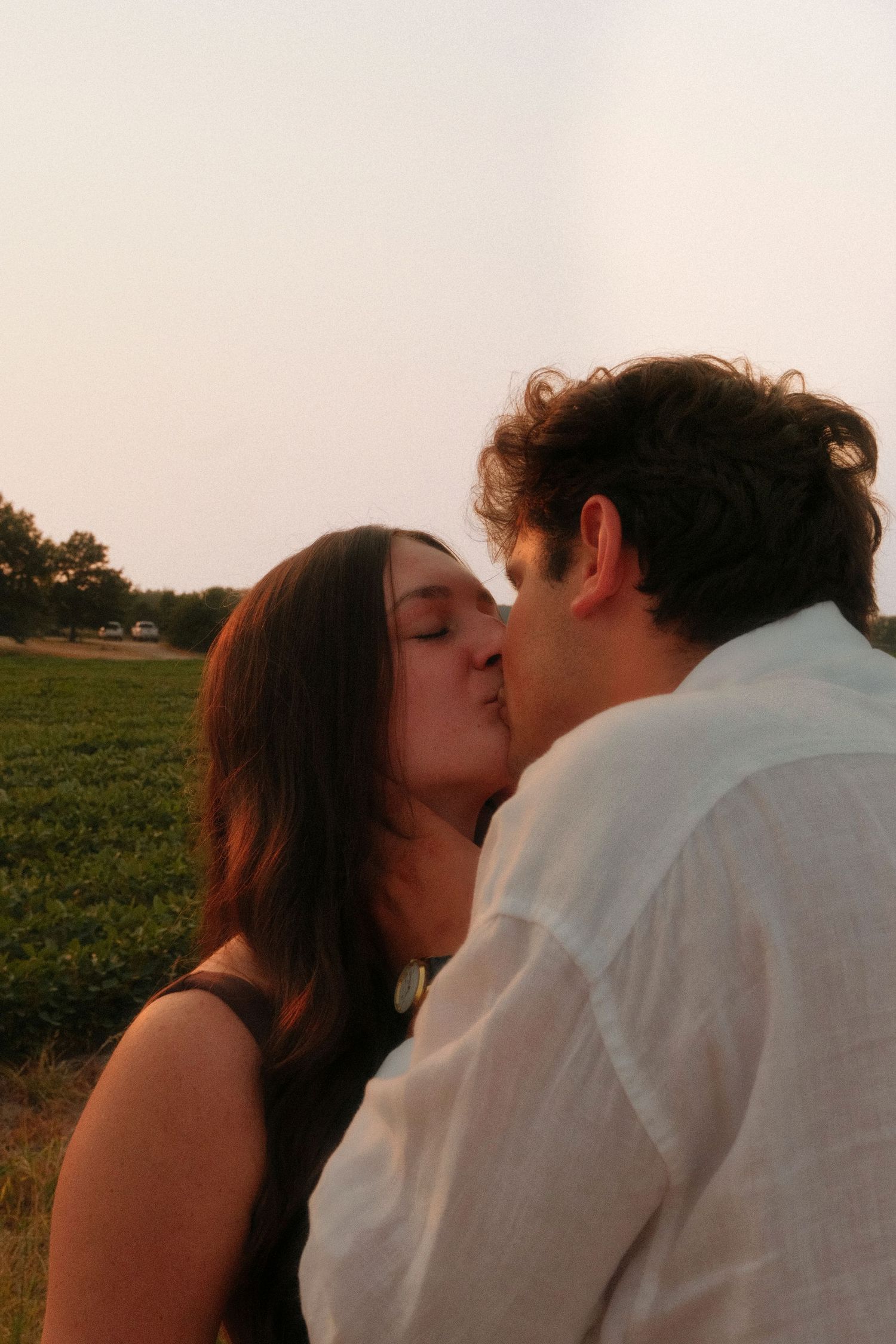 A romantic couple shares a tender kiss outdoors at sunset with green fields in the background.