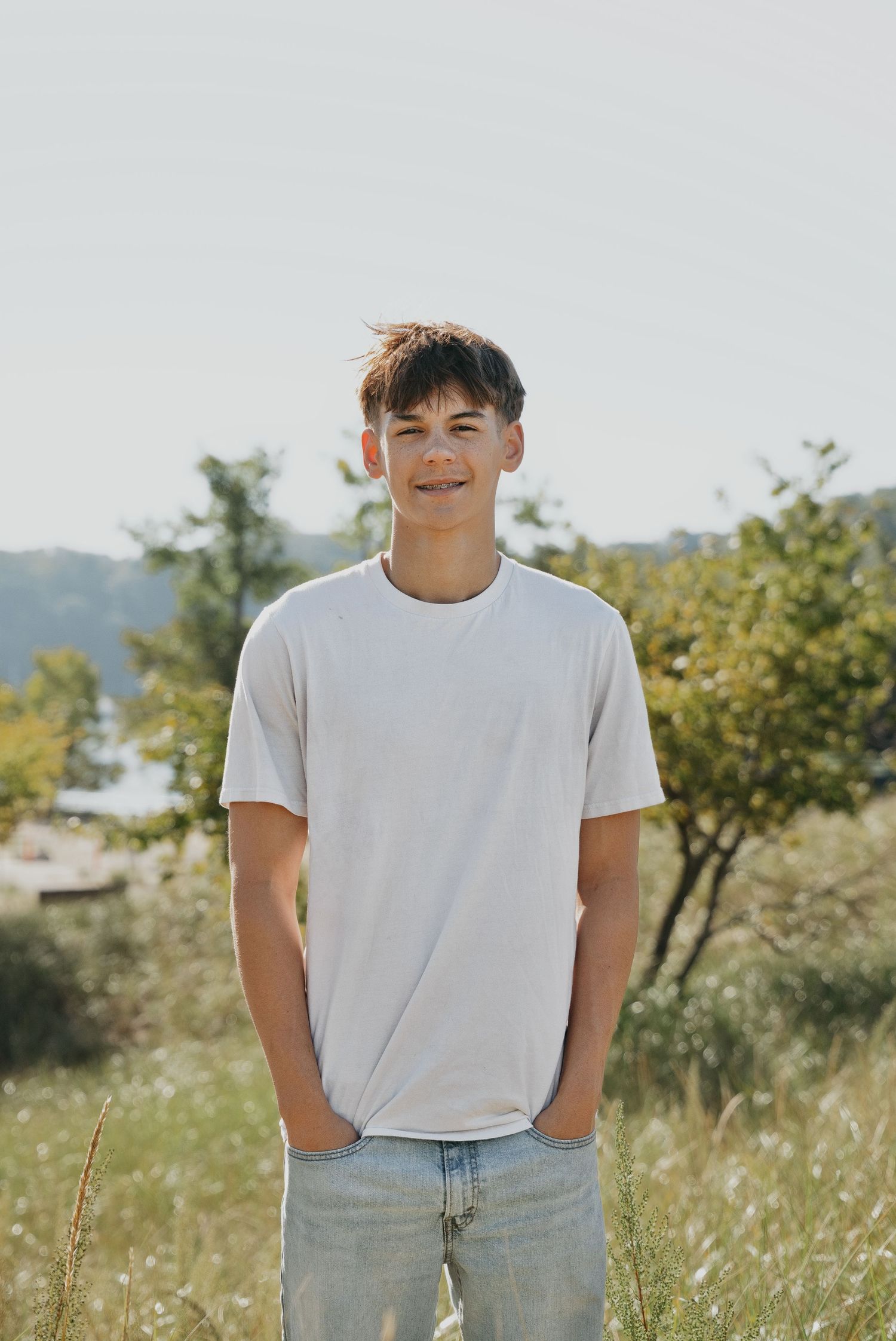 A smiling teenage boy in a white t-shirt and jeans stands outdoors in a sunny field with trees in the background.