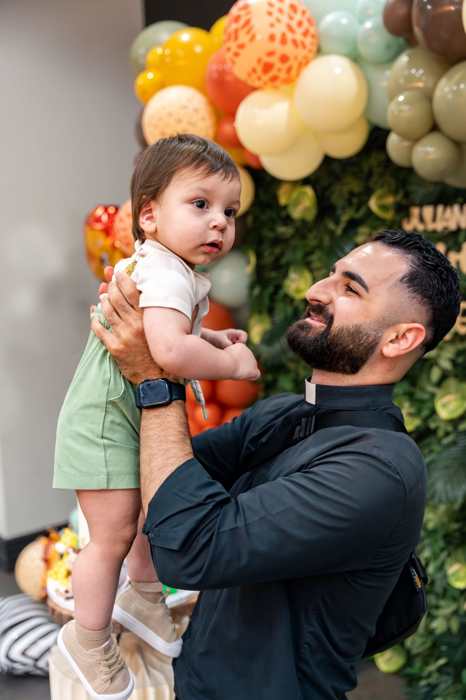 priest holding baby up in front of jungle themed backdrop