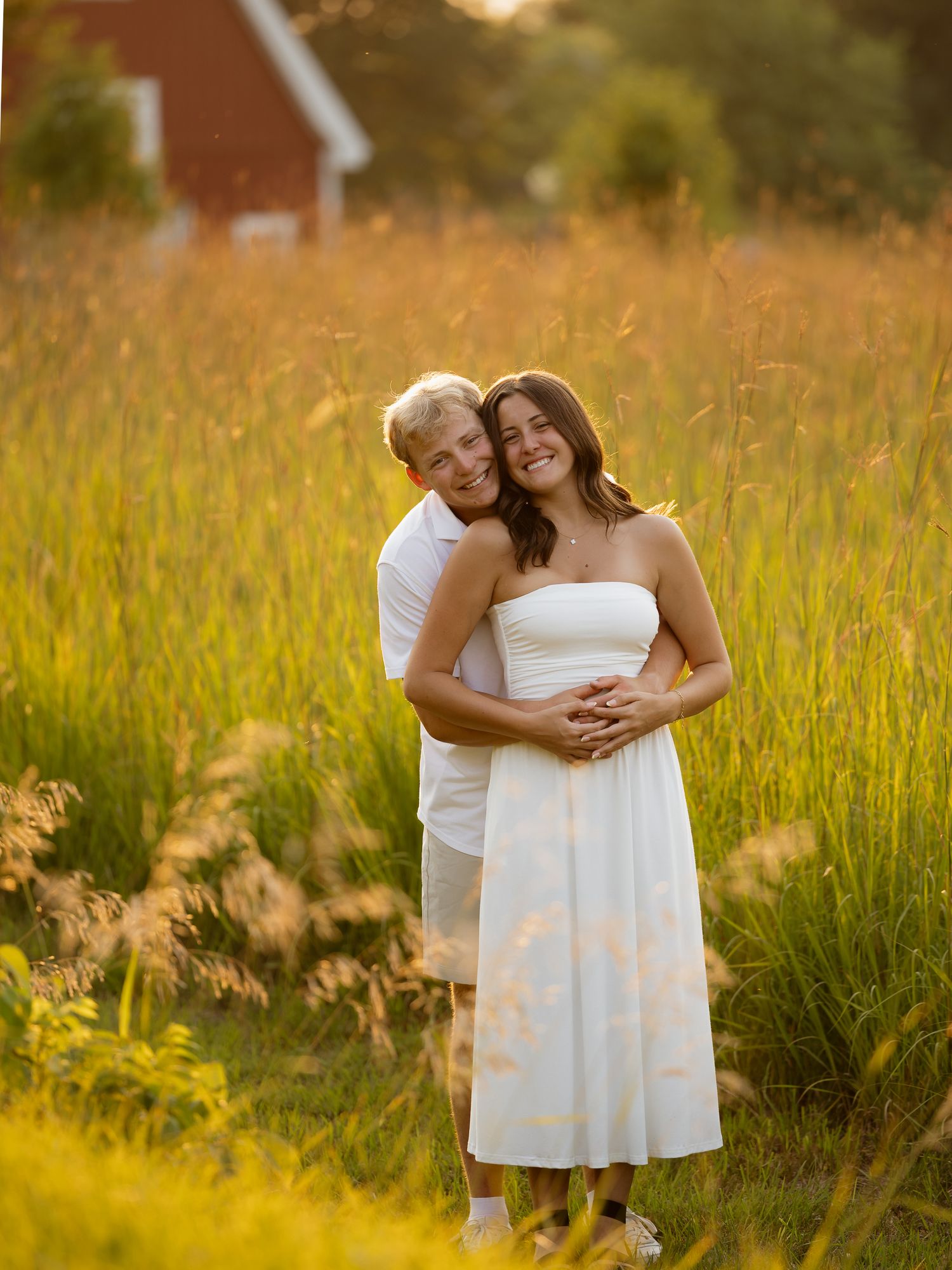 Couple embracing in golden field at sunset, woman in white strapless dress, man in white shirt, red barn background.