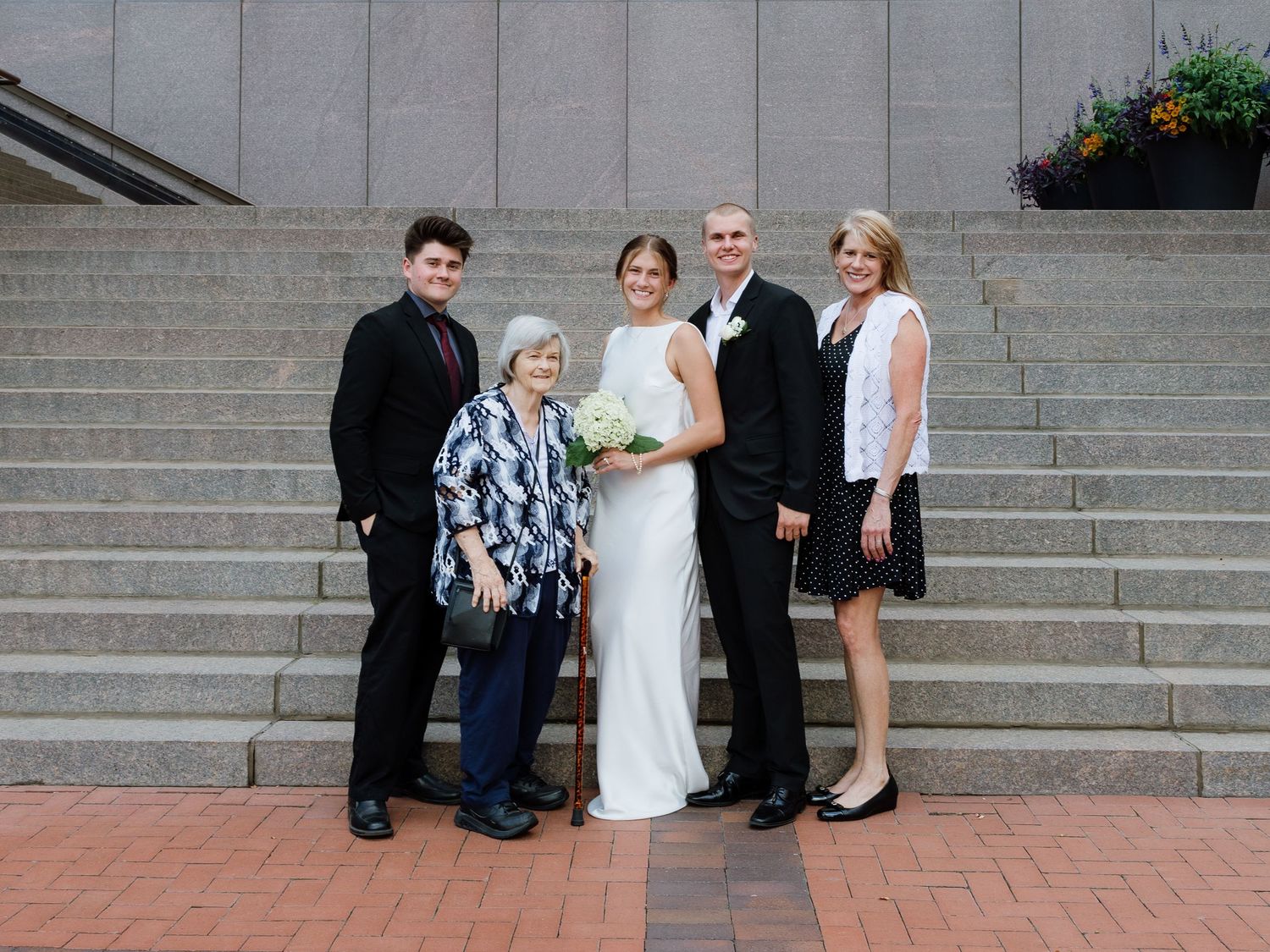 Wedding group photo of bride in white gown with bouquet, groom in suit, and three family members on brick plaza steps.