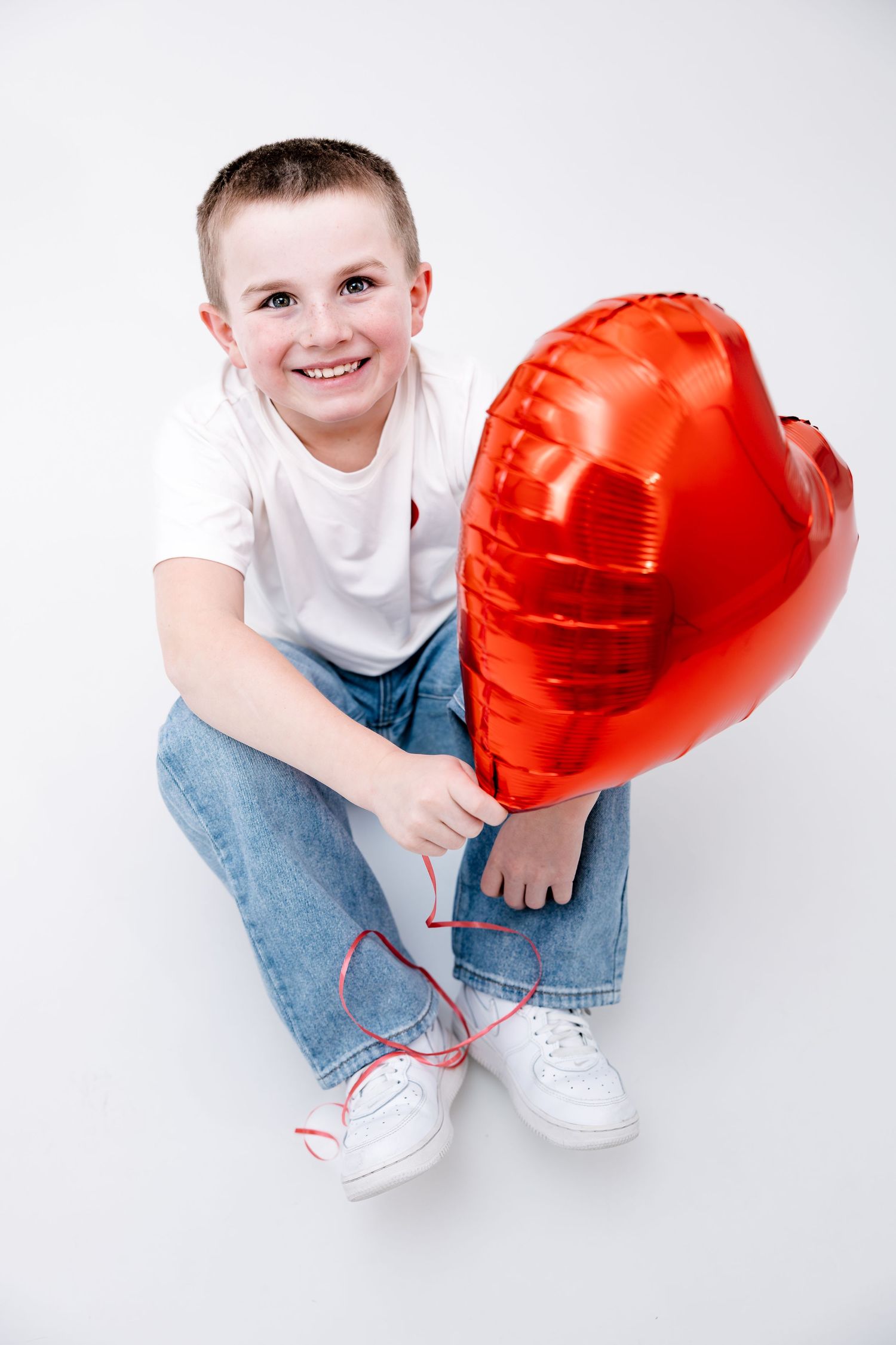 Smiling boy sitting on white floor holding a red heart-shaped balloon cheerfully.