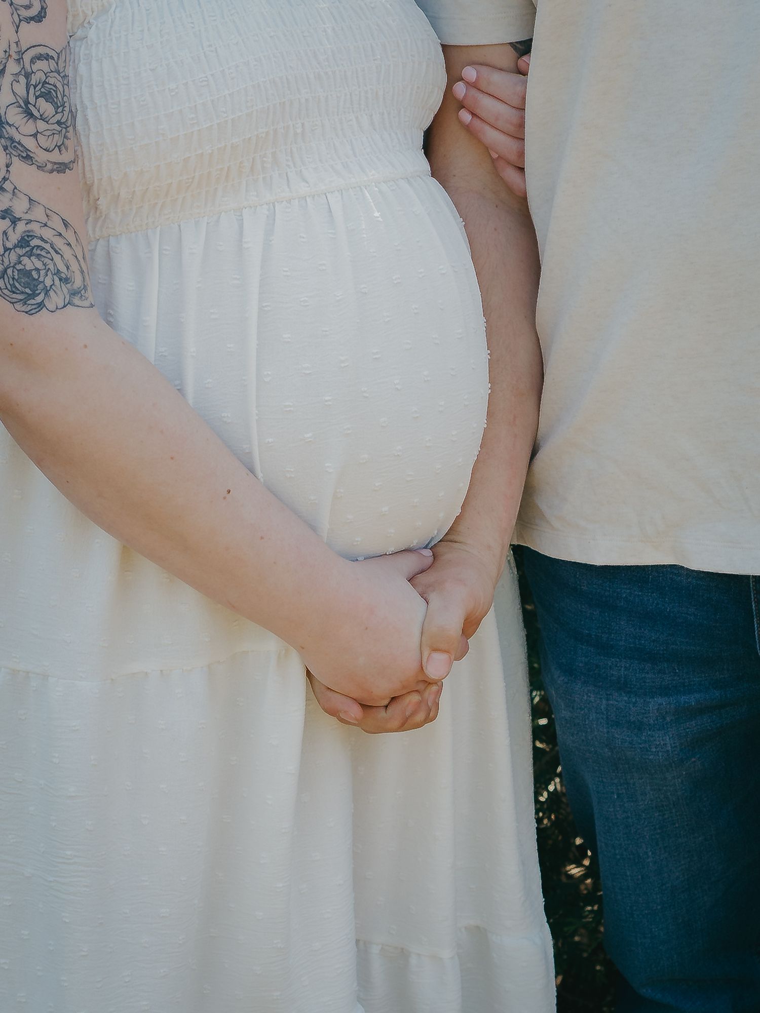 A pregnant woman in a white dress holds hands with her partner, showcasing her baby bump.