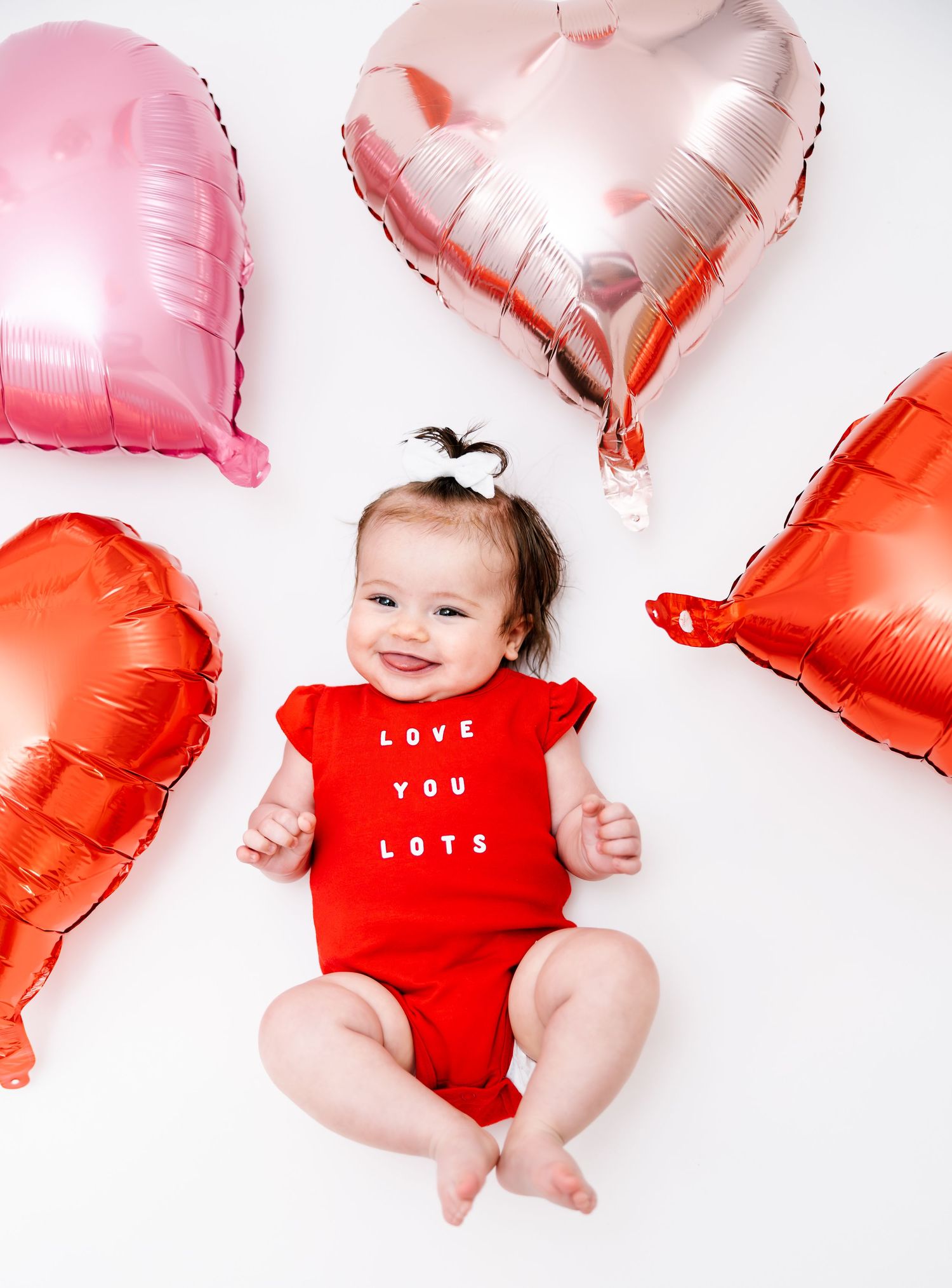 Happy smiling baby in red Love You Lots onesie surrounded by pink, orange, and red heart-shaped foil balloons.