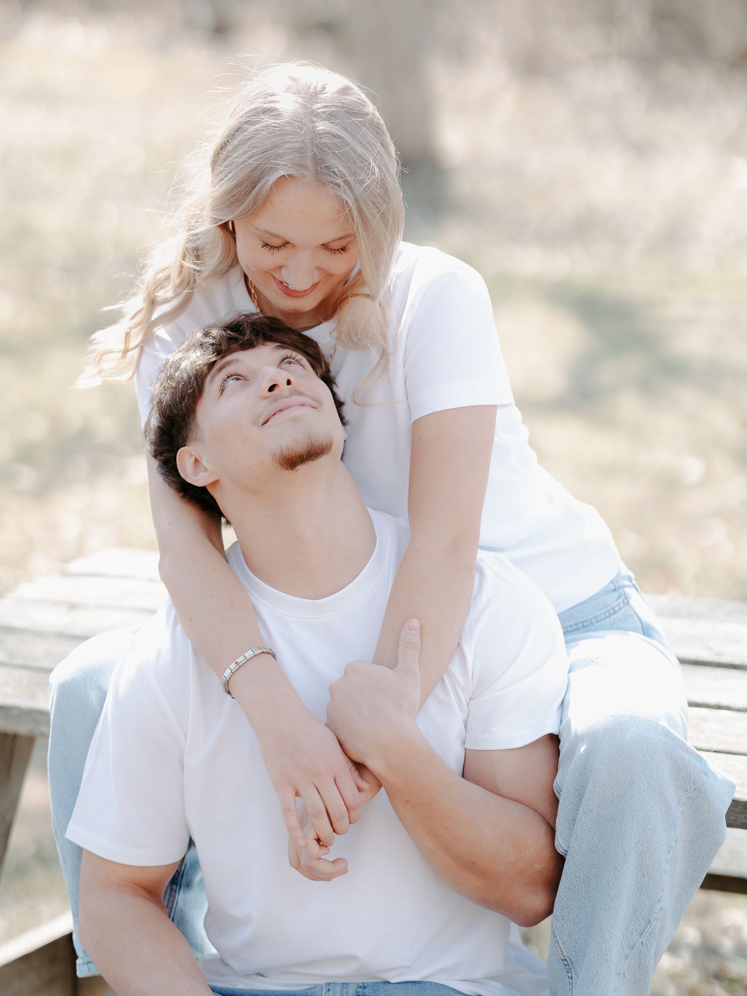 A couple in matching white t-shirts share a tender moment outdoors, woman embracing man from behind on a wooden bench.