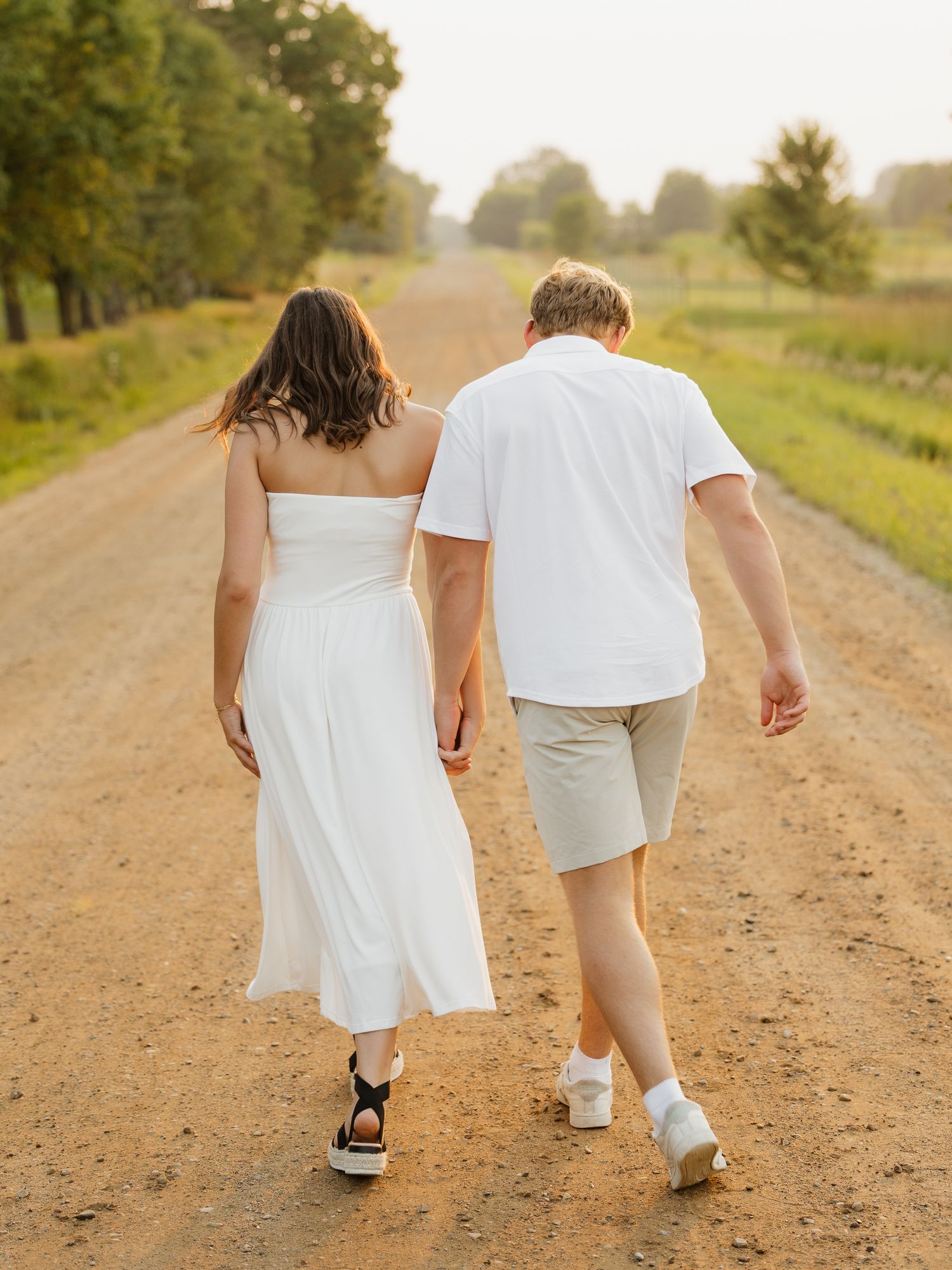 Couple walking hand in hand down a scenic dirt road, woman in white dress and man in white shirt and khaki shorts.