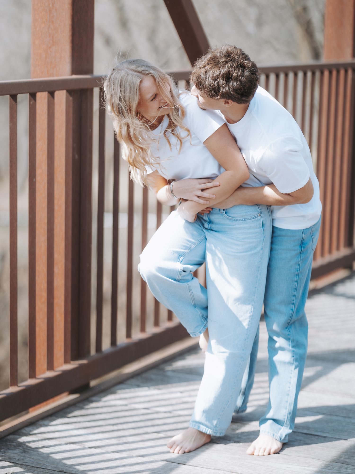 Young couple in white shirts and jeans playfully embracing on a sunny wooden bridge.