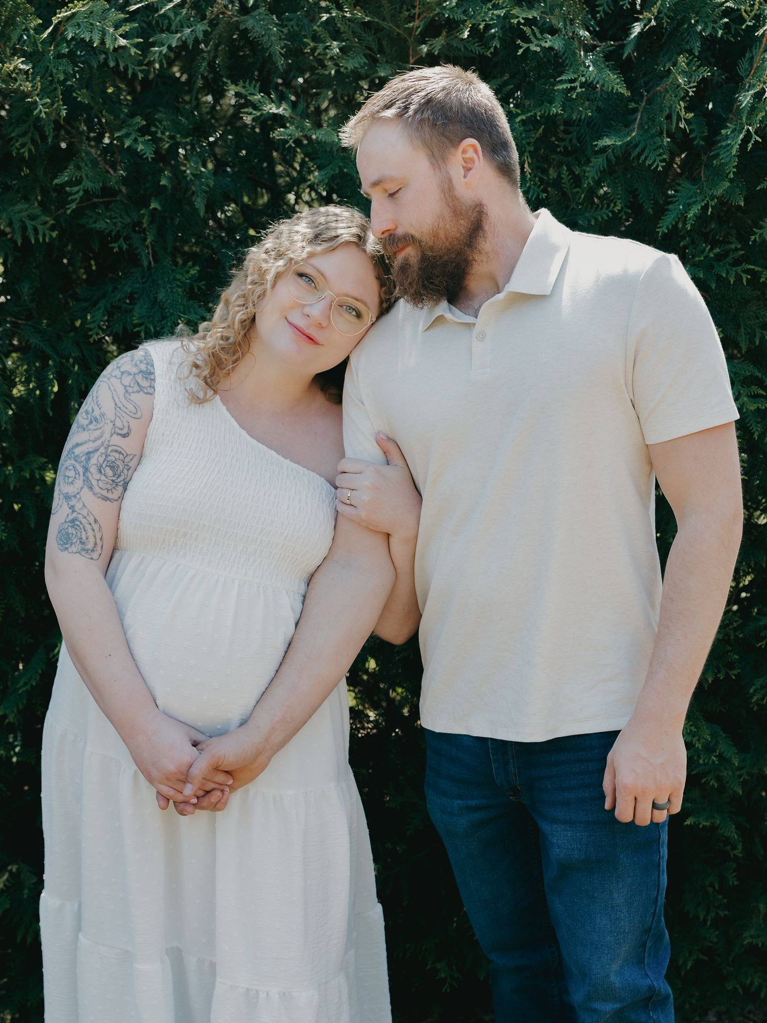 A pregnant couple stands together in front of green trees, the woman in a white dress leaning on the man.